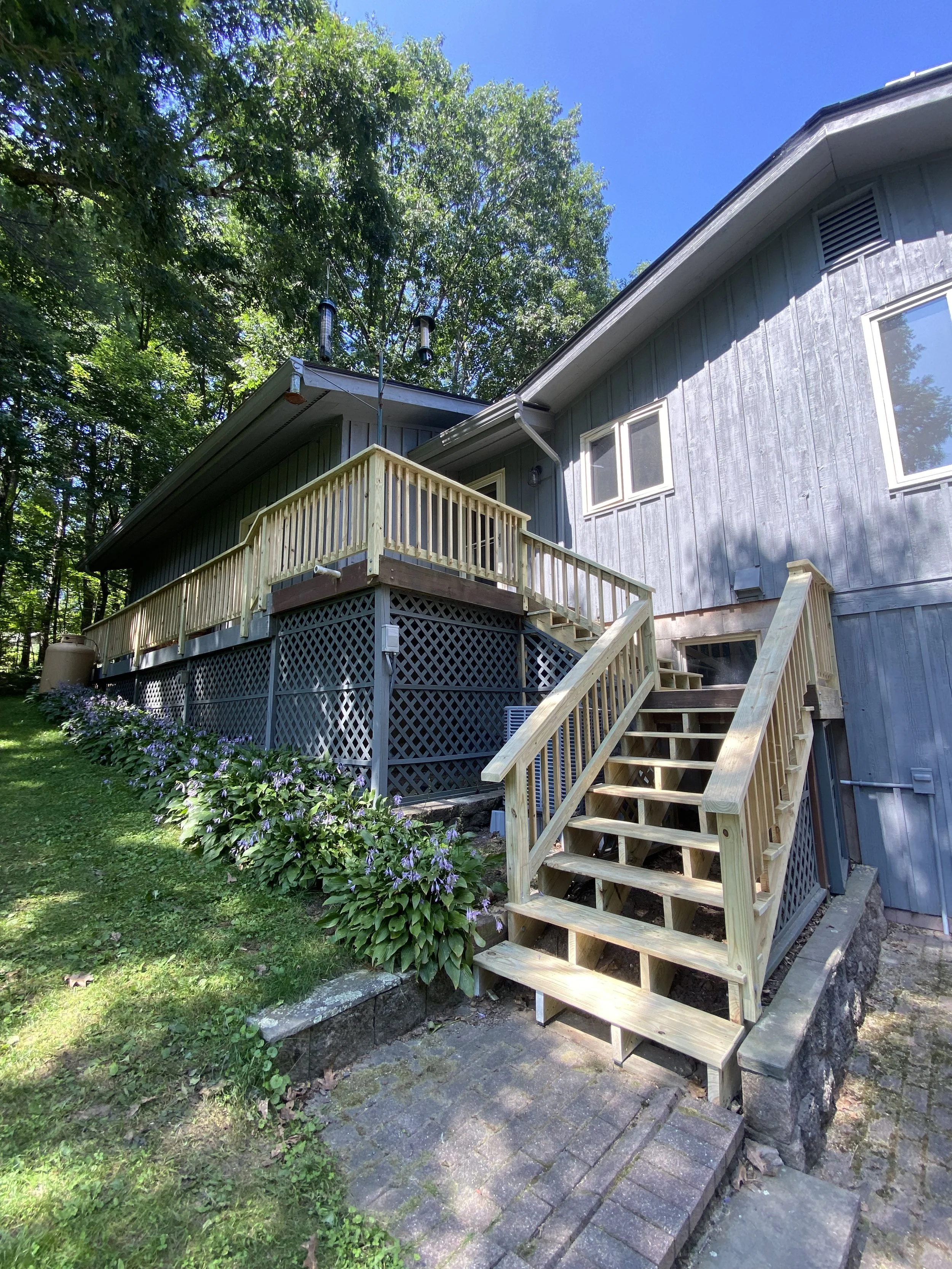 A wooden staircase leads up to a house with a deck surrounded by lush greenery and tall trees, with a clear blue sky overhead.