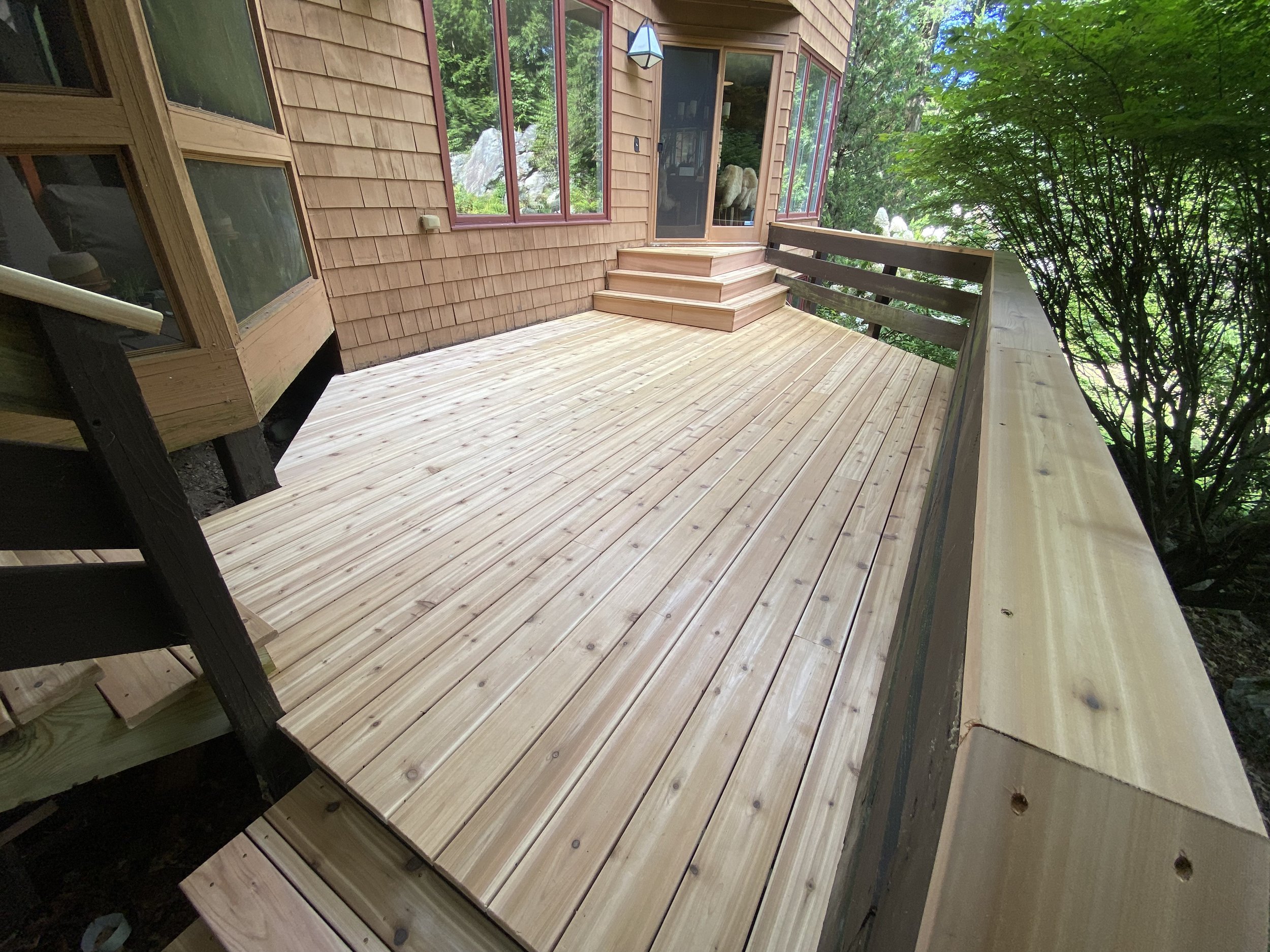 A newly built wooden deck attached to a house with stairs leading up to a glass door, surrounded by green trees.