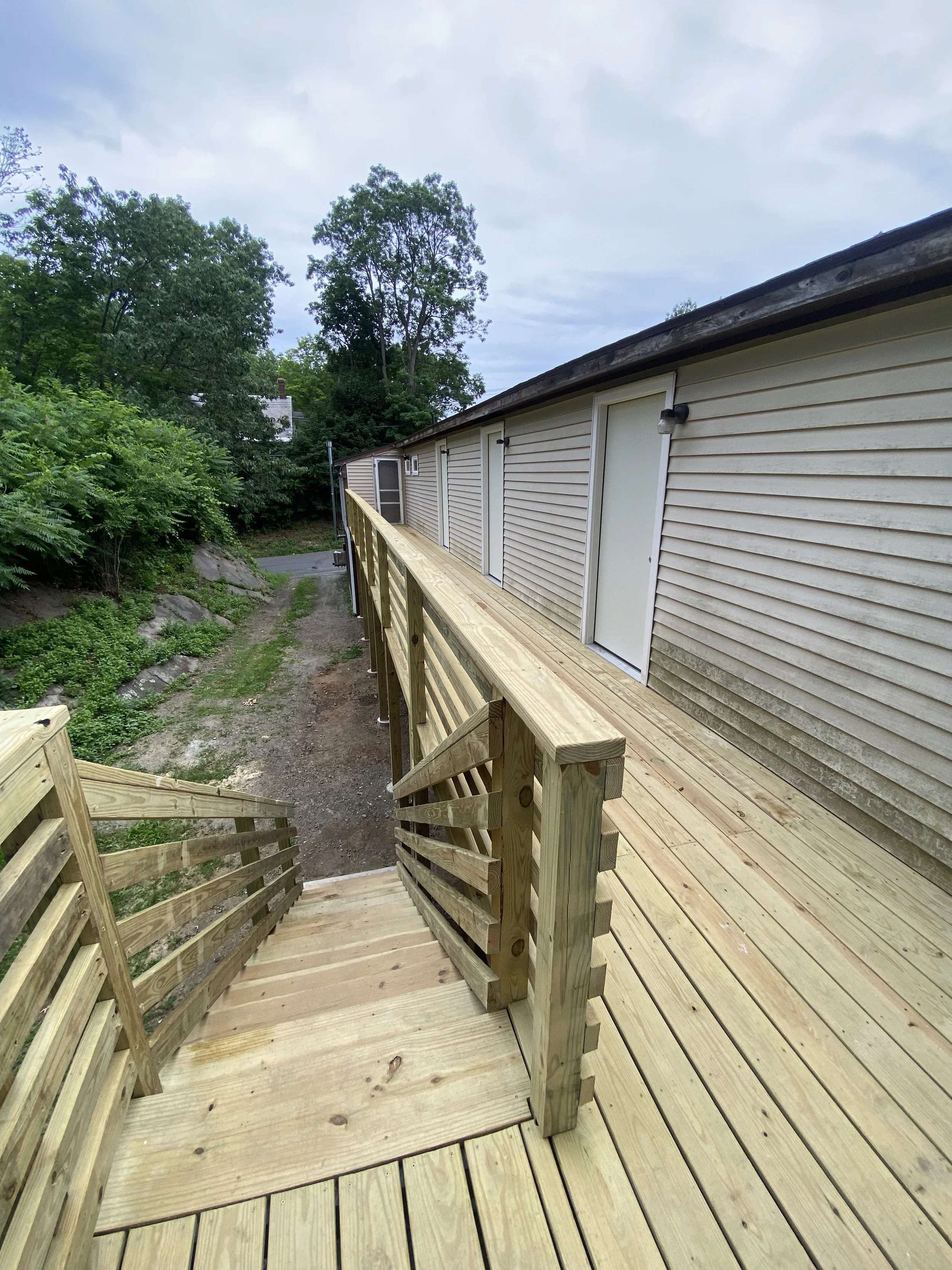 Wooden deck with stairs and railing attached to a beige house with a door, overlooking a dirt path and green trees.