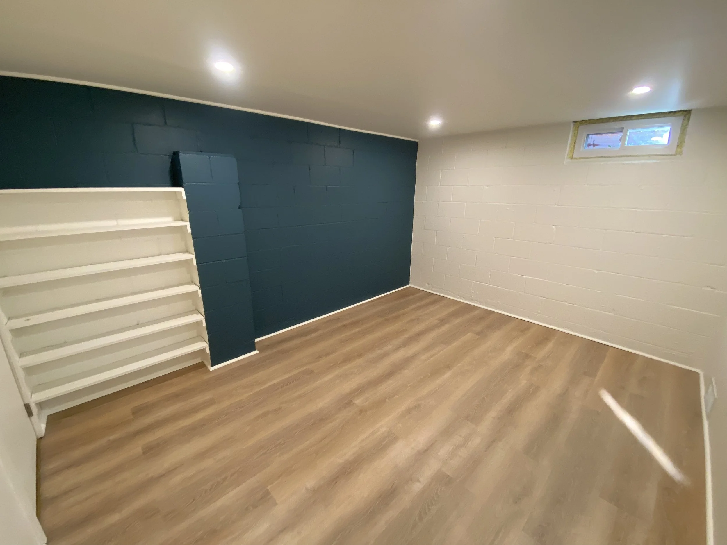 Empty basement room with white brick wall, blue painted wall, small window, ceiling lights, and wooden floor.