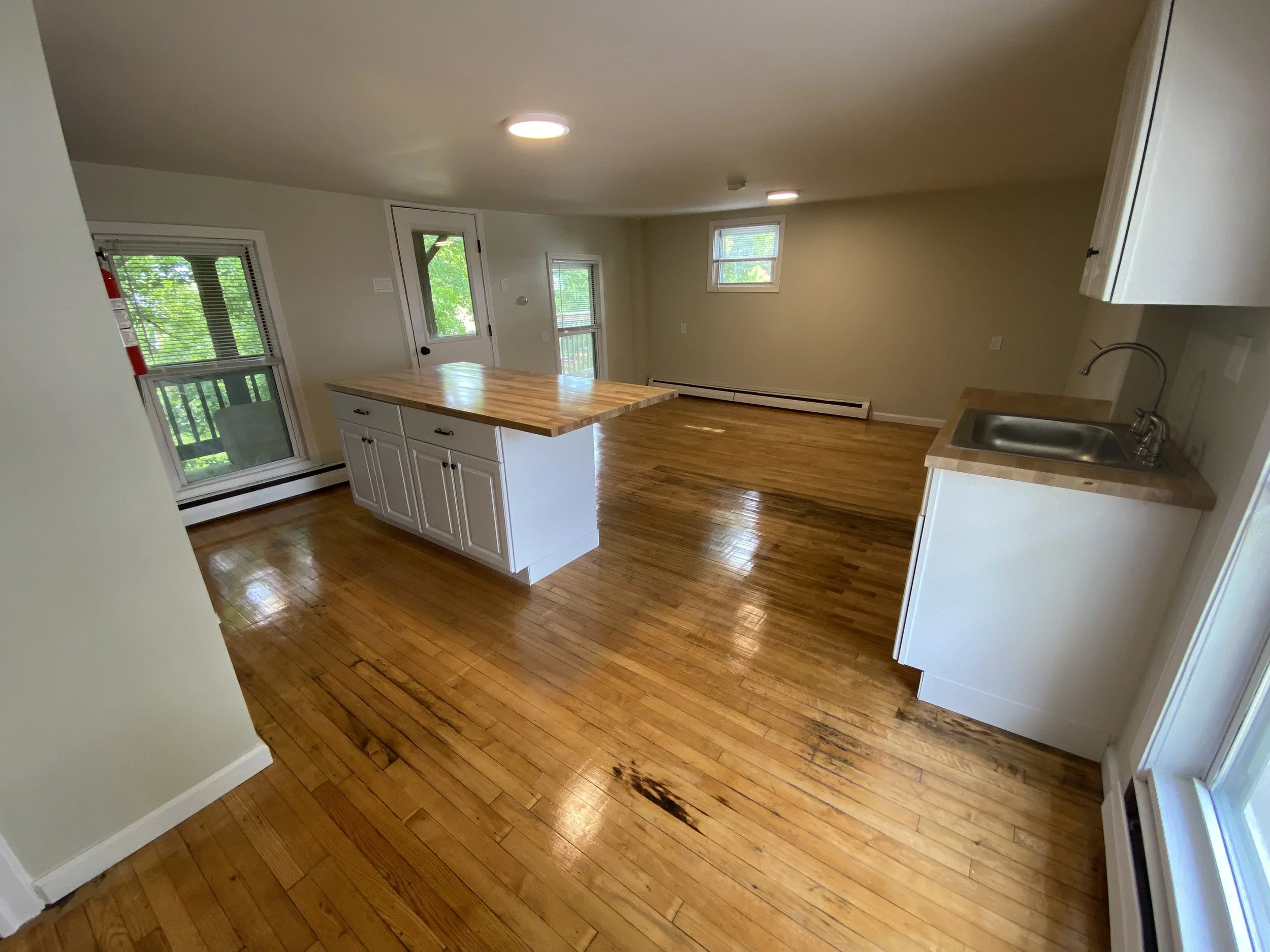 Empty kitchen and living area with hardwood floors, white cabinets, a small kitchen island, a sink, windows, and a door leading to a balcony.