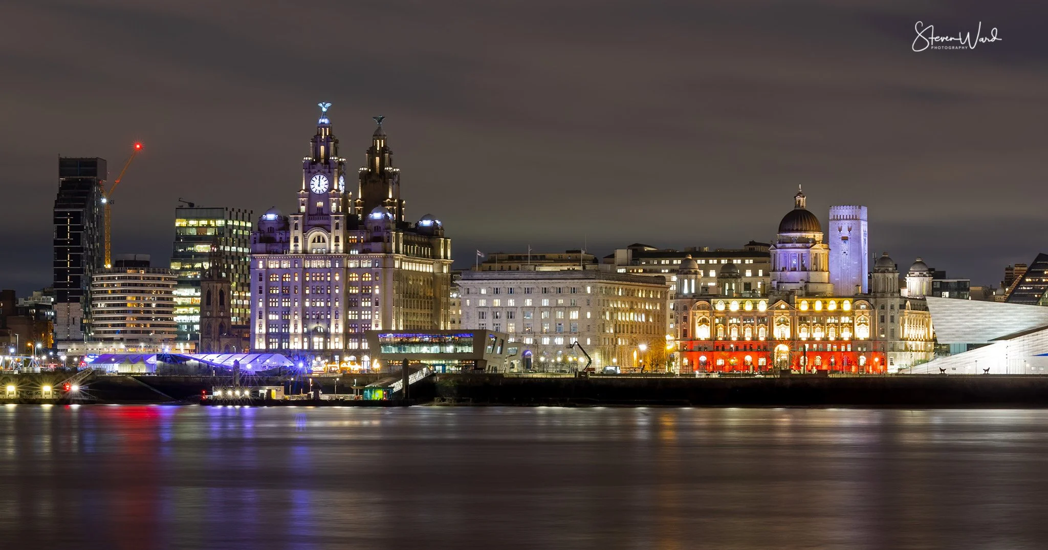 Nighttime cityscape of Liverpool, England, with illuminated historic buildings and modern skyscrapers along the waterfront. Reflections on the water are visible in the foreground.
