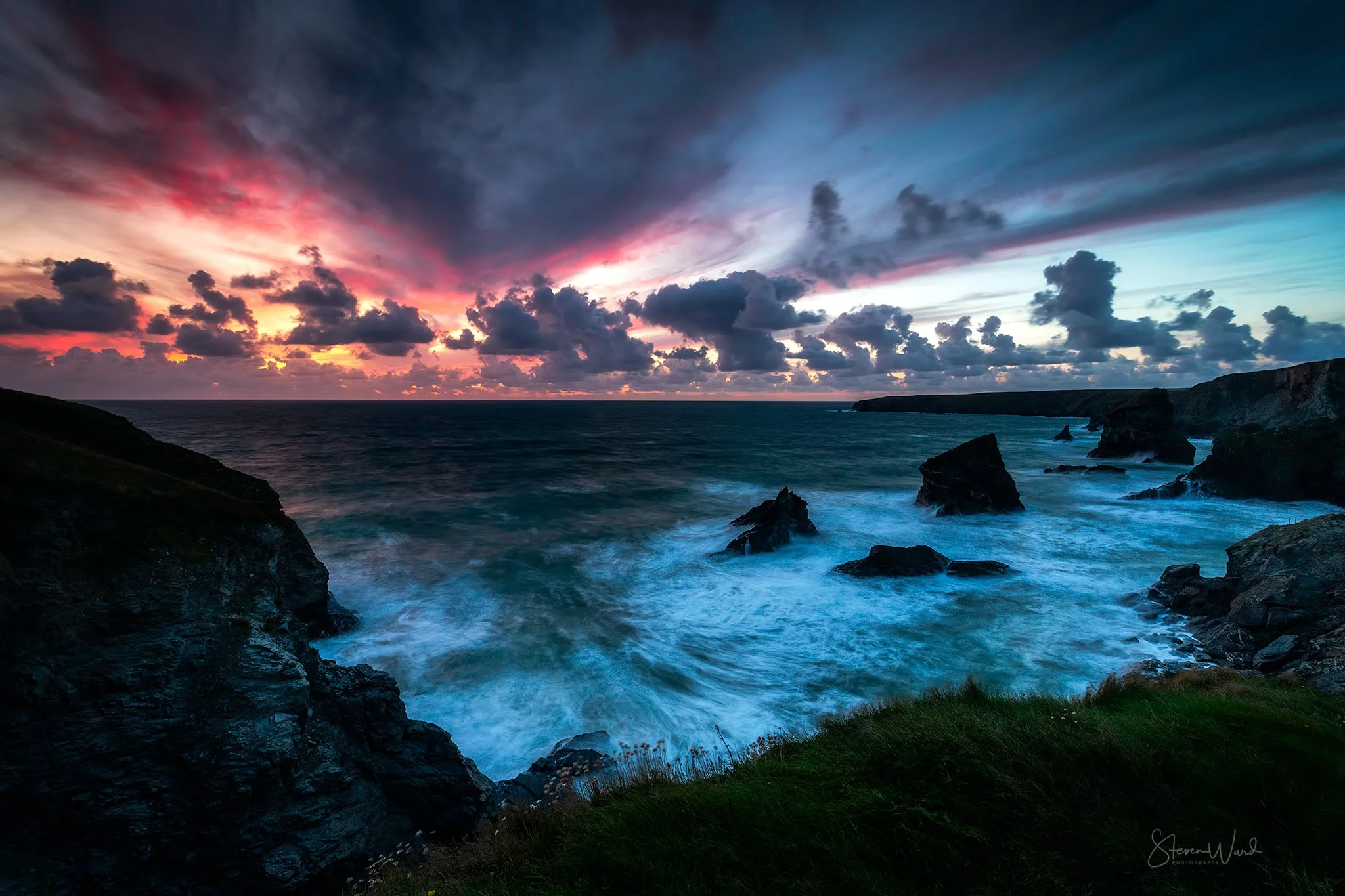 A dramatic sunset over a rugged coastline with dark clouds, churning ocean waves, and rocky outcroppings.
