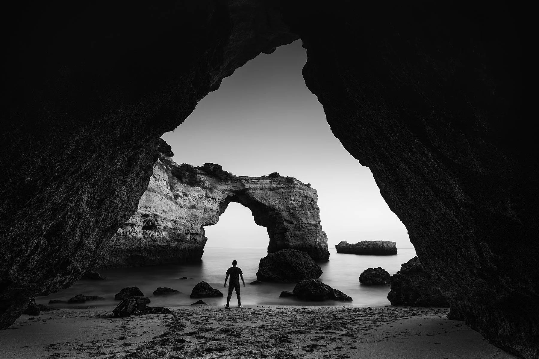 A person standing on a beach framed by large dark rock formations with a natural arch in the background, black and white photo.