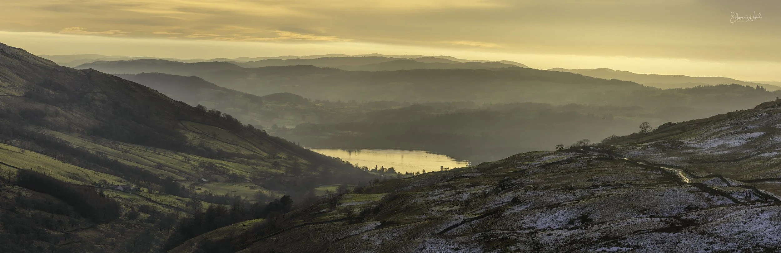 A scenic view of rolling hills and mountains during sunset or sunrise, with a river or lake reflecting the sky, some snow on the ground, and scattered trees and fields.