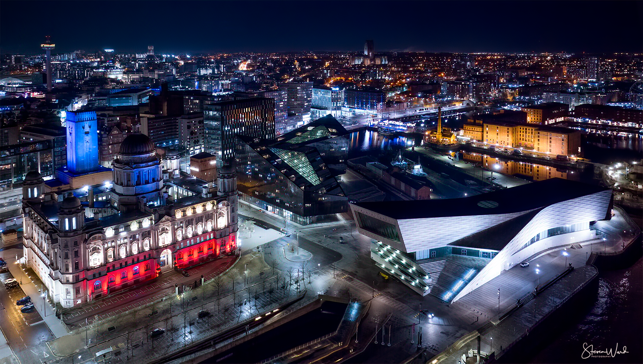 Night aerial view of a cityscape featuring a historic building with illuminated red and white lights, and a modern, uniquely shaped building with white and gray exterior lights, surrounded by parking lots and roads, with numerous lit-up skyscrapers i