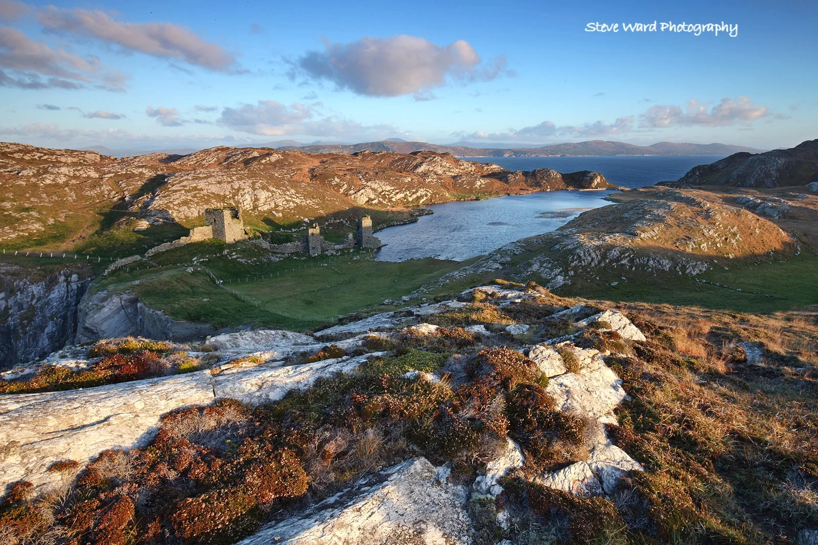 Scenic landscape of a rocky hillside overlooking a body of water with ruins of old stone structures, surrounded by hills under a partly cloudy sky at sunset.
