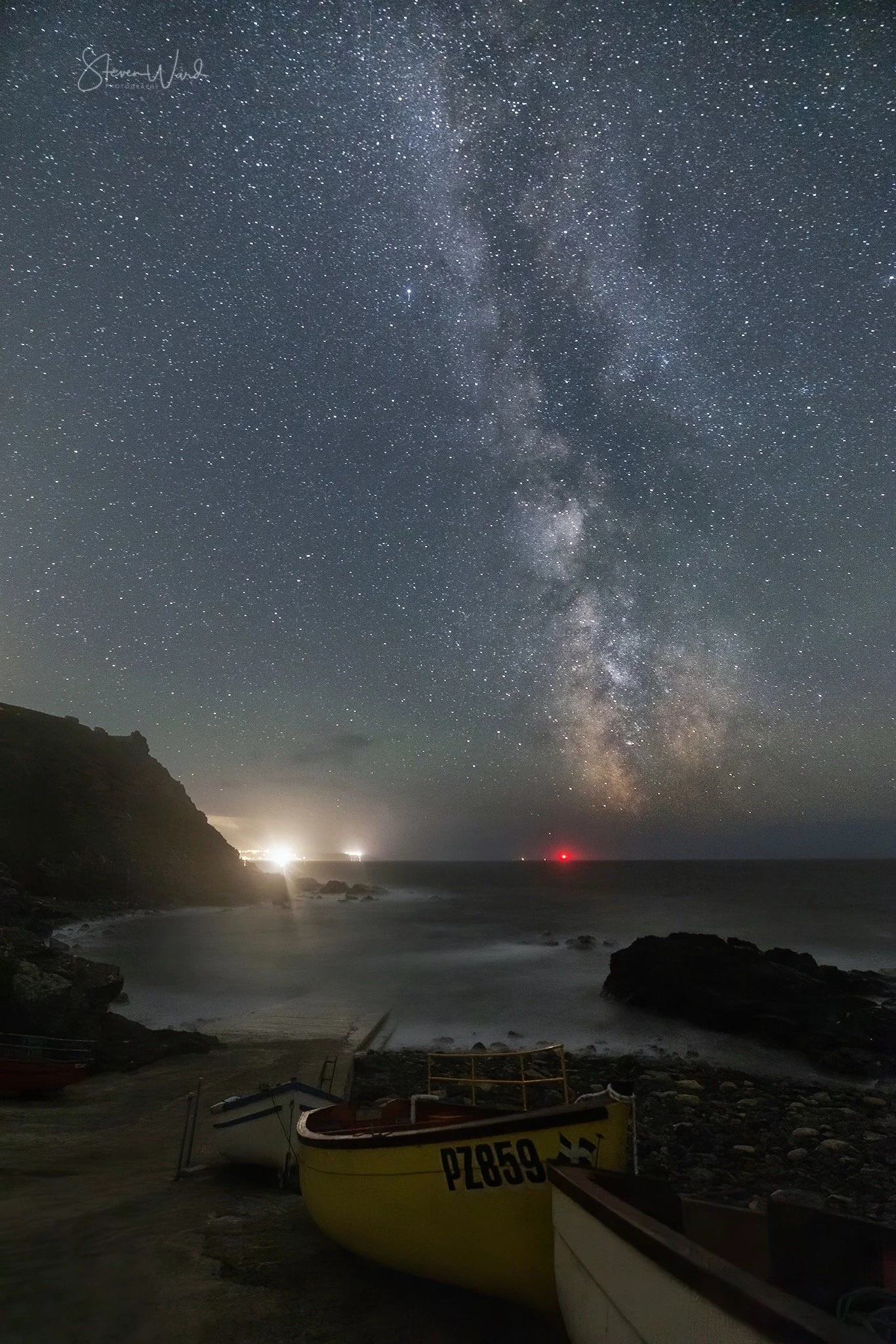 Nighttime scene of a starry sky, including the Milky Way galaxy, over a rocky coastline with boats on the shore and a distant lighthouse