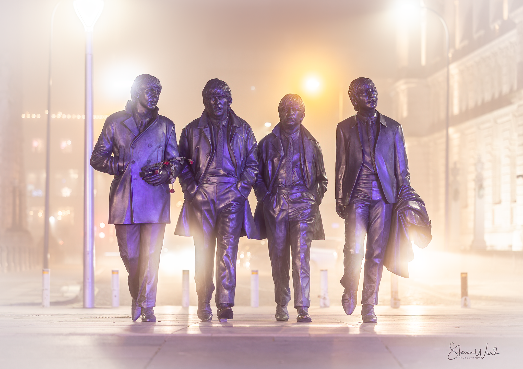 Bronze statue of The Beatles walking on a city street at night, with streetlights and buildings in the background.
