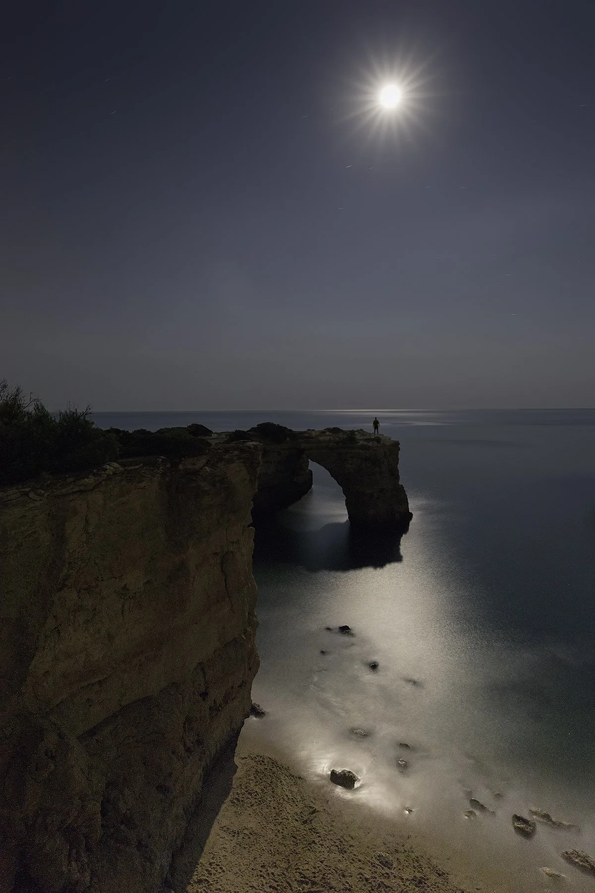 Night view of a rocky coastal landscape illuminated by moonlight, featuring a natural rock arch formation and a person standing on top of the arch, with calm ocean waters below and a bright full moon in the sky.