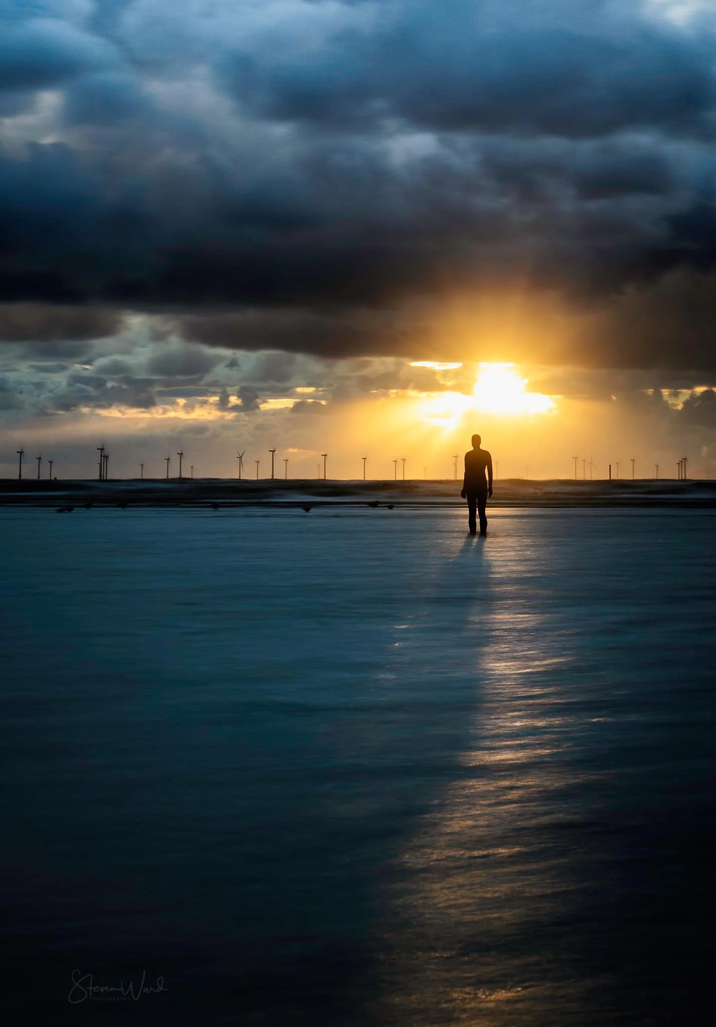 A person standing alone on a beach during sunset, with dark clouds overhead and wind turbines in the distance.