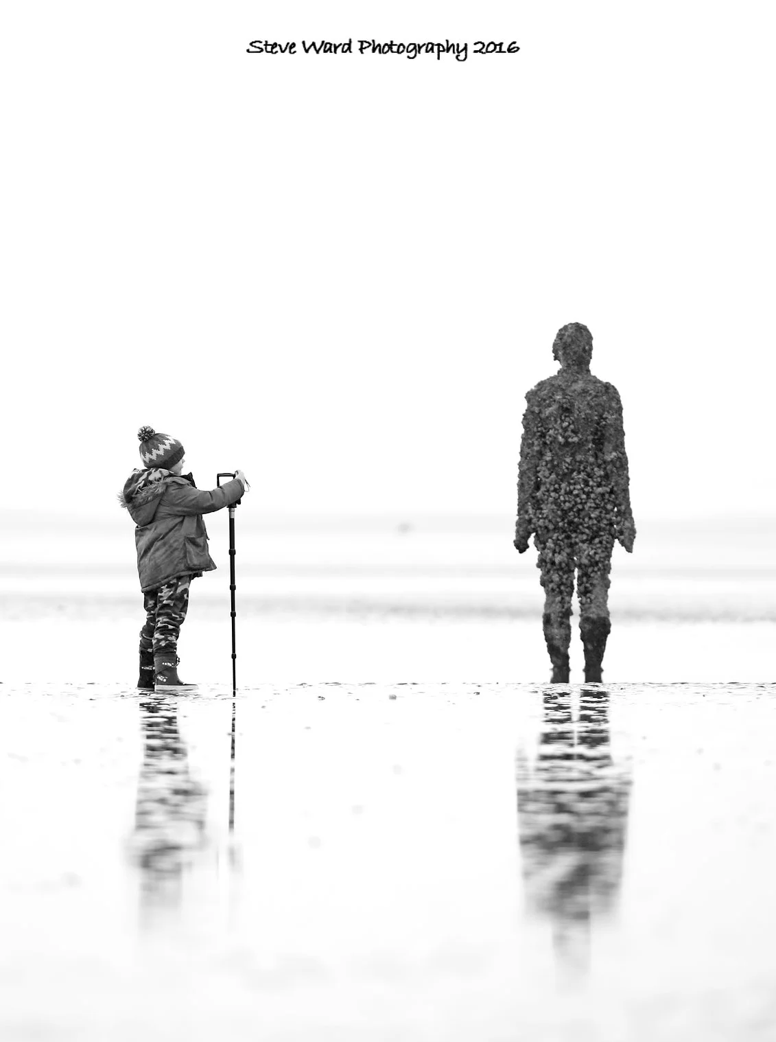 A child in winter clothing taking a photograph of a person covered in flowers, ambos standing in shallow water with reflections, scene in black and white, titled 'Steve Ward Photography 2016'.