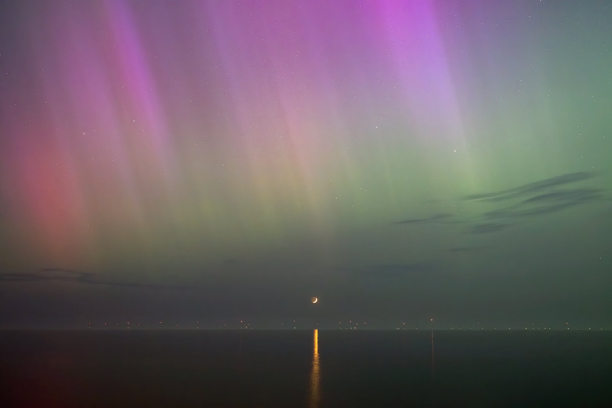 Northern lights in the sky over the ocean with a crescent moon and reflection on the water.