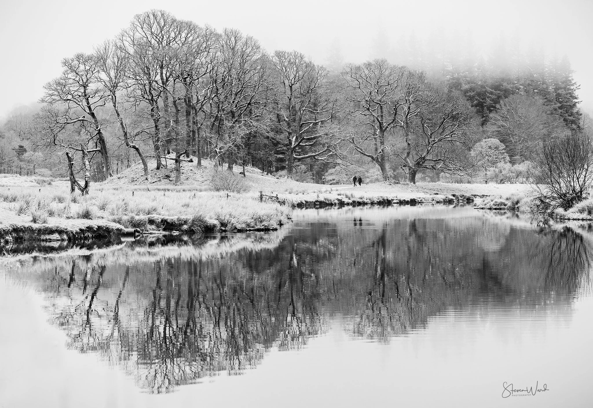 A black and white photo of a snowy winter landscape featuring leafless trees along the banks of a calm river, with snow-covered ground and misty background.