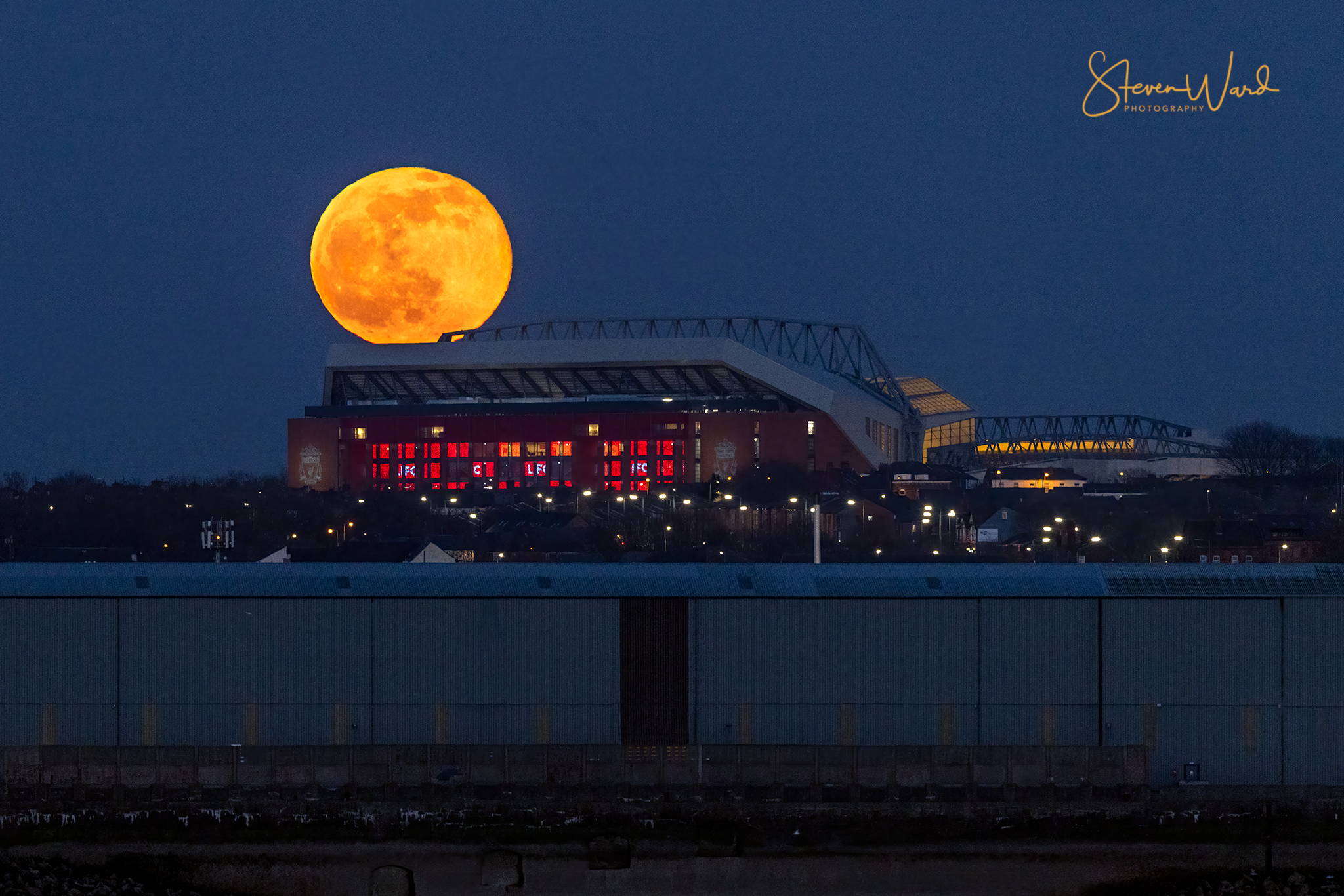 A large orange full moon rising behind a stadium with illuminated windows and a blue sky.