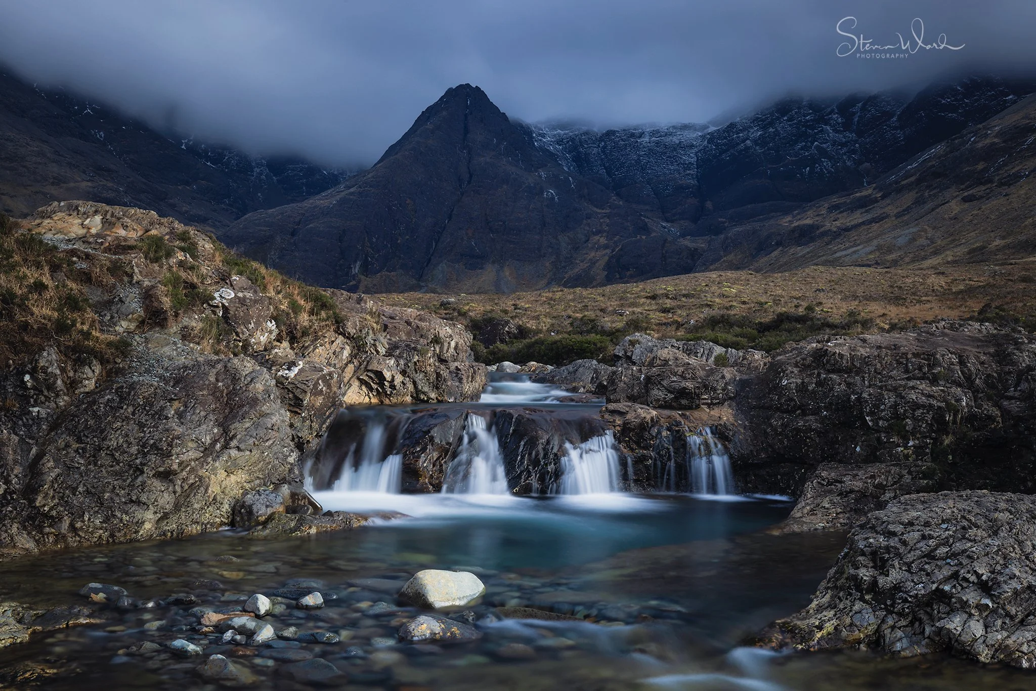 Mountains with dark clouds, flowing stream over rocks in a valley.