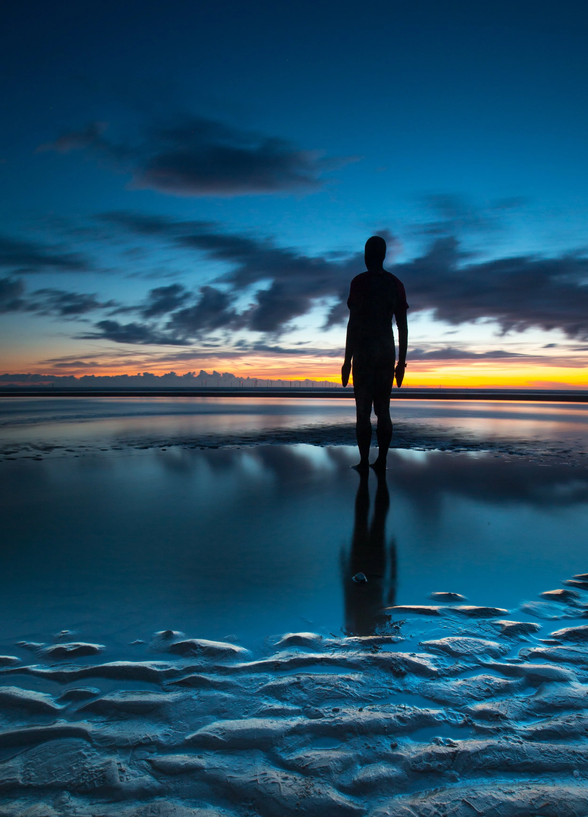 Silhouette of a person standing on the beach at sunset with reflecting water, dark clouds in the sky, and a colorful horizon.