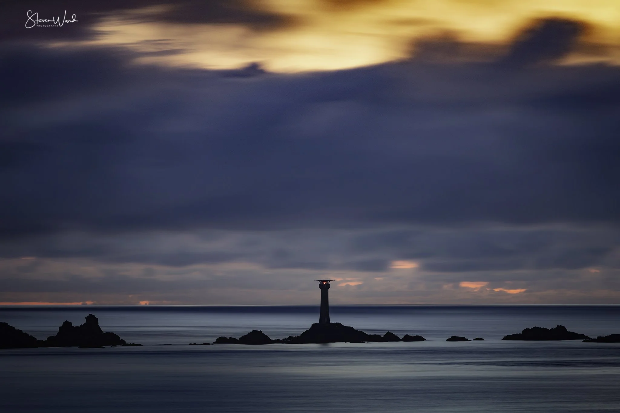 A lighthouse on a rocky island surrounded by calm ocean waters at sunset, with dark storm clouds and a colorful sky above.