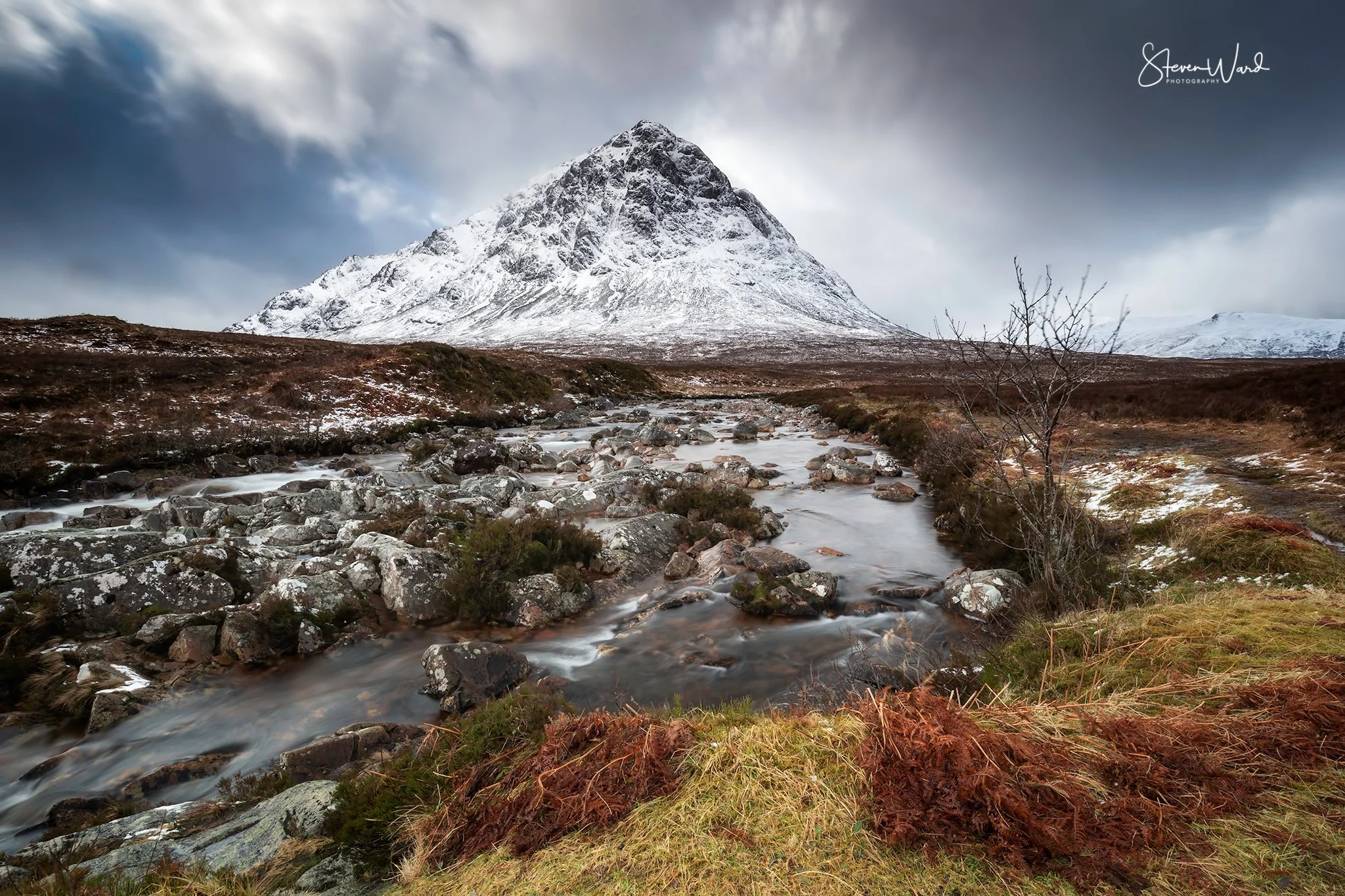 Snow-capped mountain with cloudy sky, flowing stream with rocks in foreground, and brown and green plants on the riverbank.