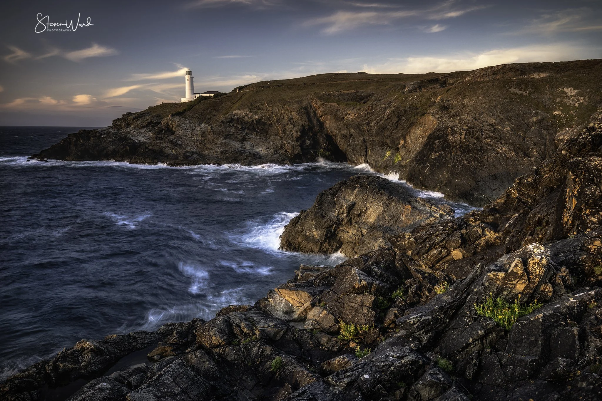Coastal scene with rocky shoreline, ocean waves, and a lighthouse on a hill at sunset.