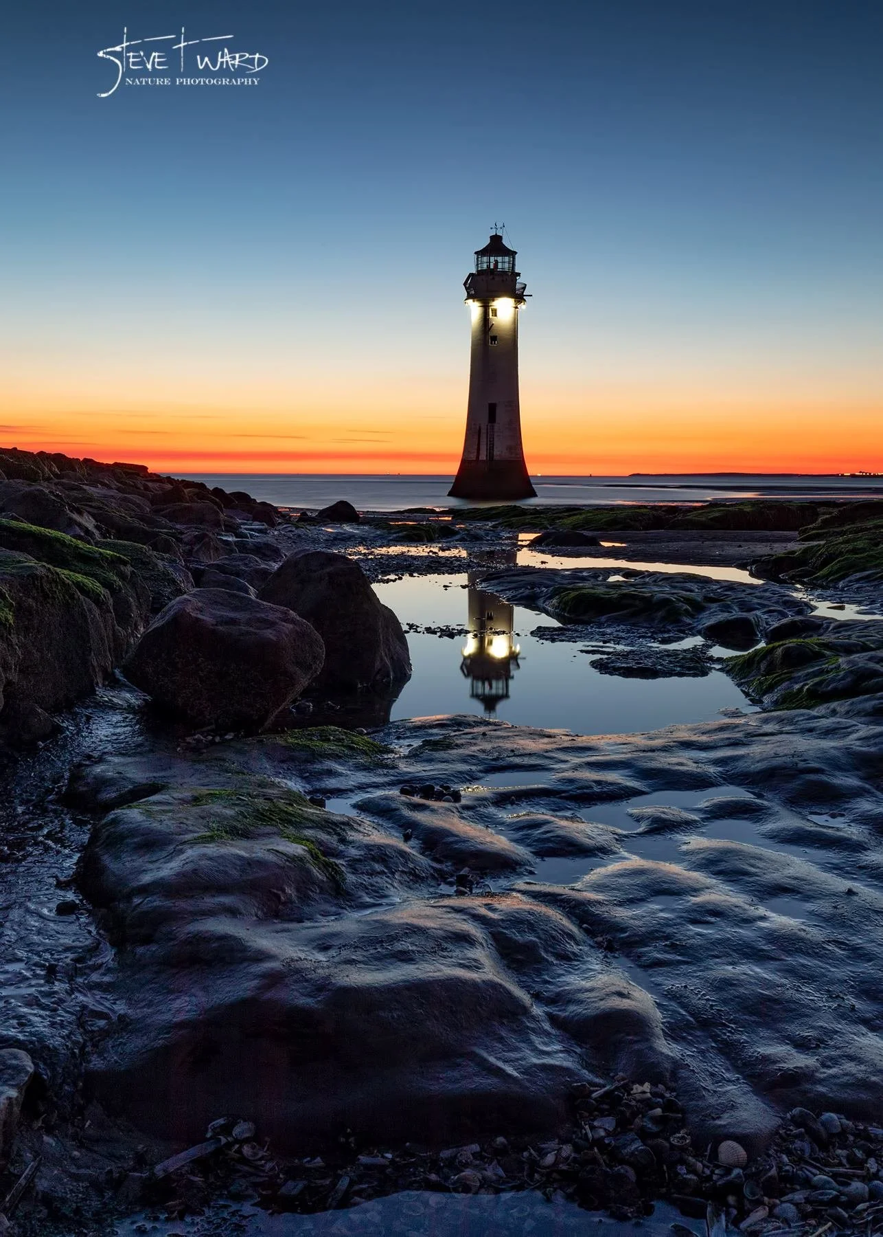 A lighthouse standing tall by the rocky shoreline during sunset, with its reflection visible in nearby puddles and calm water, featuring an orange and blue sky.