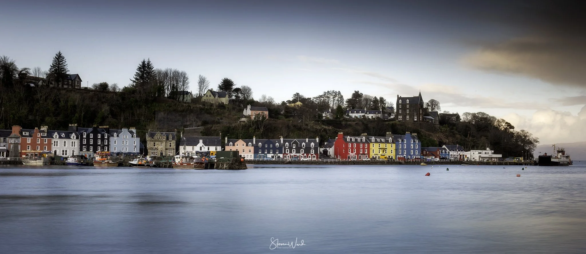 Colorful houses along a waterfront with boats docked at the pier and a hill with trees and houses in the background.
