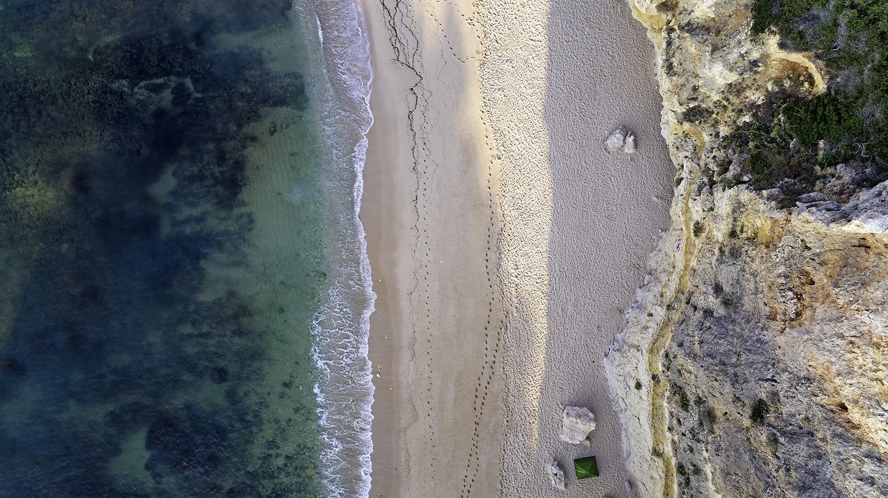 Aerial view of a beach with sandy shore, footprints, cliffside, rocks, and crystal-clear ocean water.