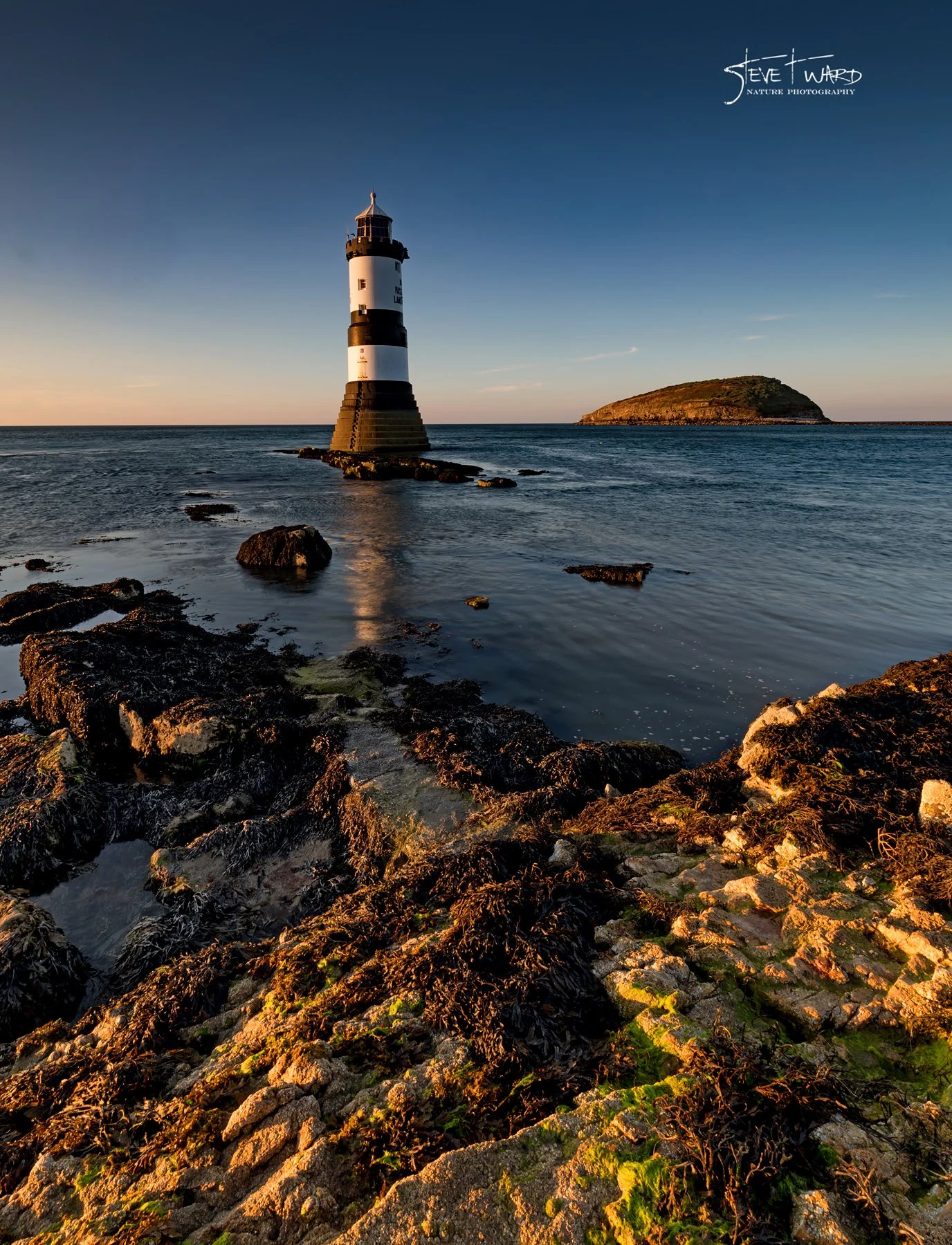 A lighthouse stands on rocks and water, with a small island in the background during sunset.