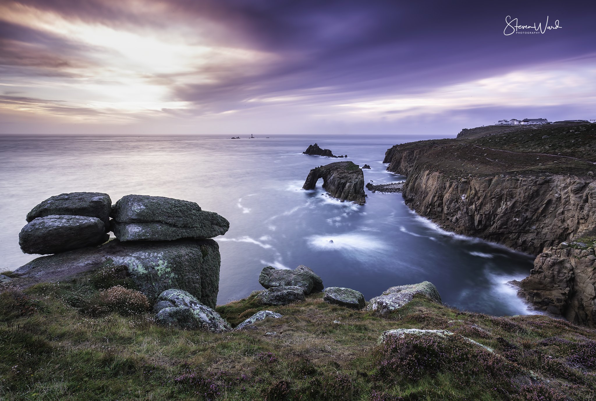 Coastal scene with rocks and cliffs at sunset, with purple and pink sky and ocean waves.