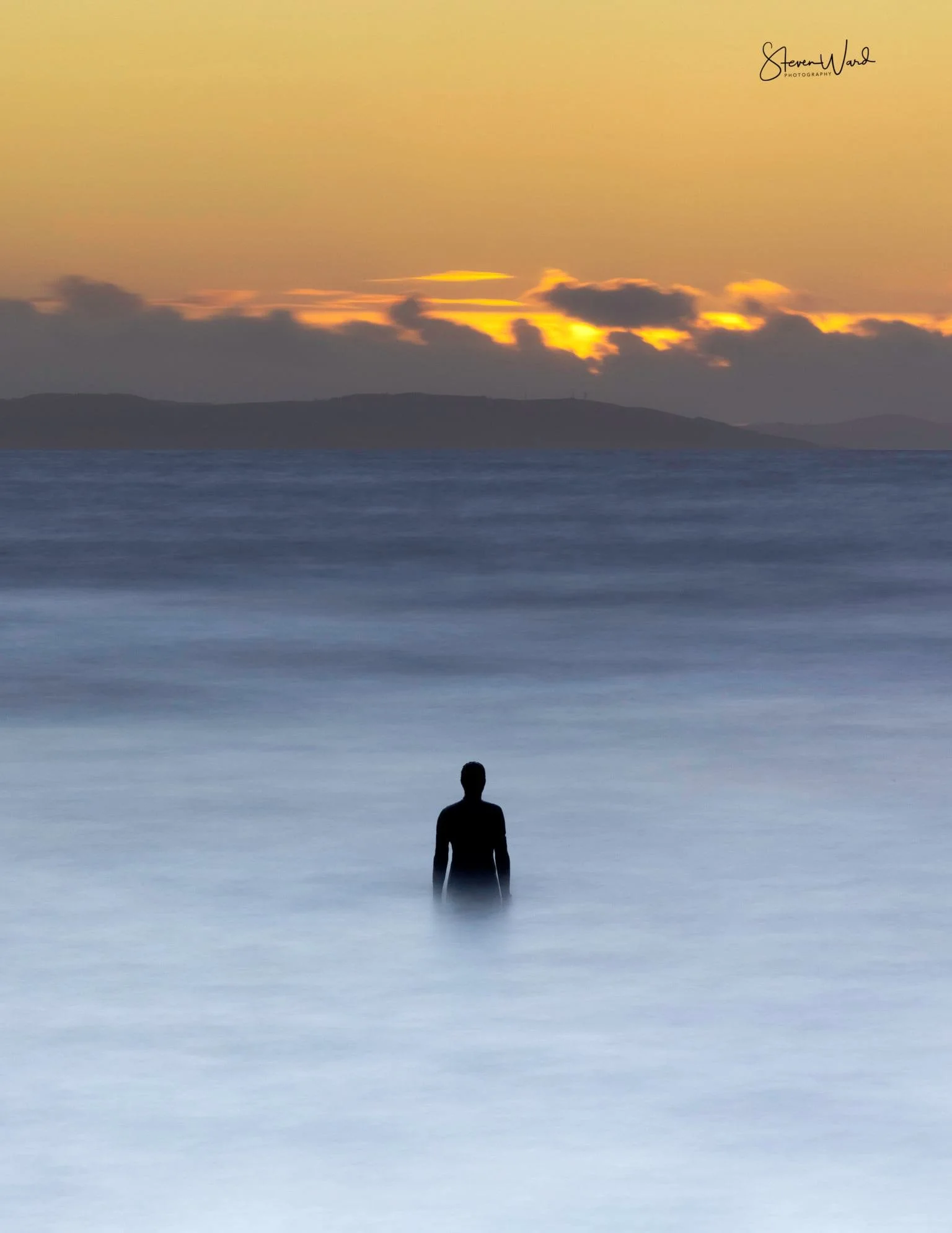Silhouette of a person standing in the ocean during sunset with yellow sky and clouds in the background.