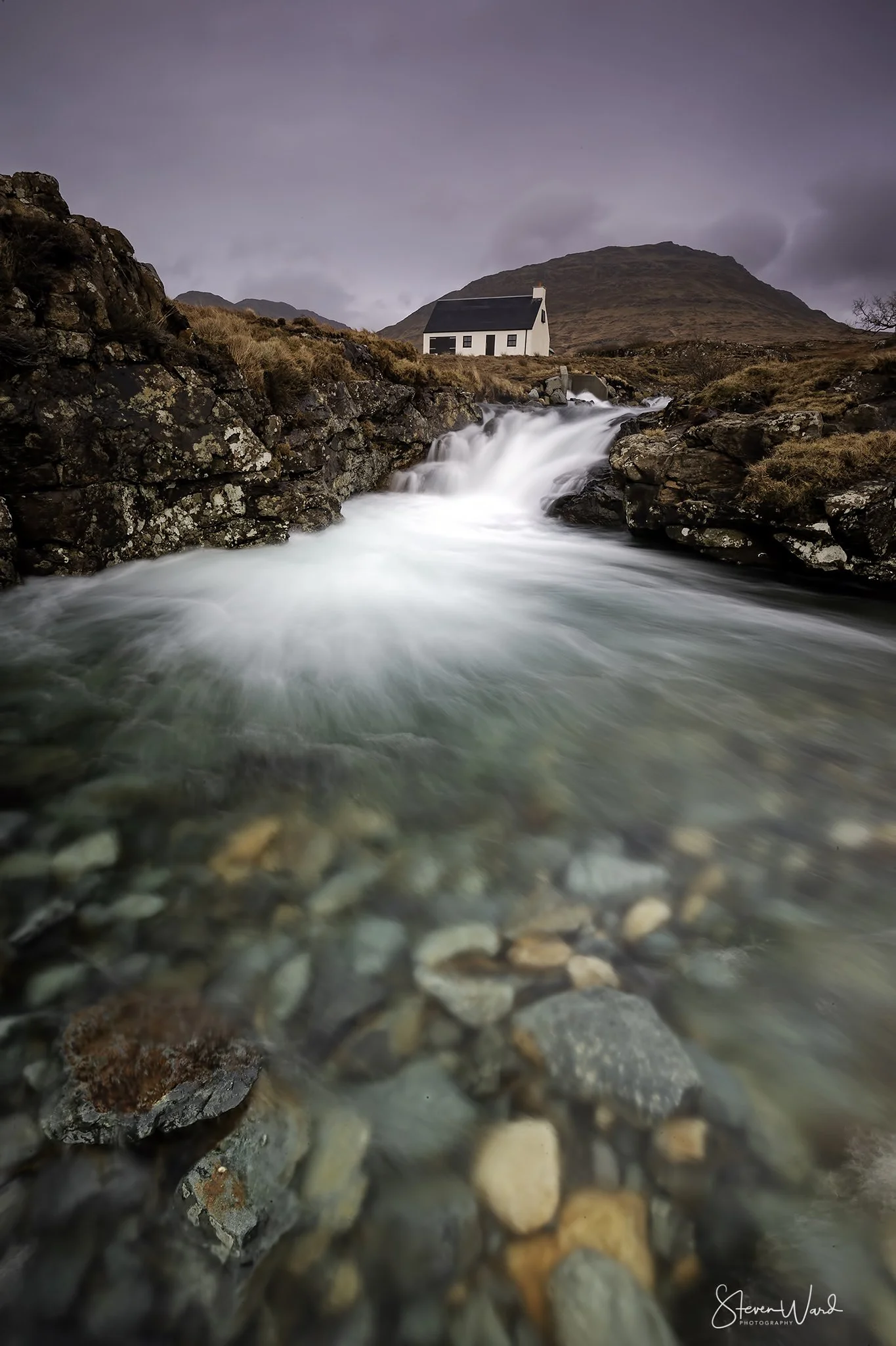 Flowing water over rocks with a white house and mountain in the background, under an overcast sky.