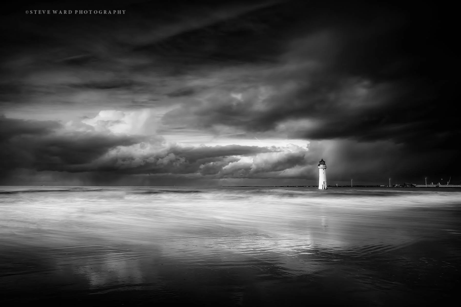 Black and white photograph of a lighthouse on the coast under a stormy sky with dark clouds and a reflective ocean surface.