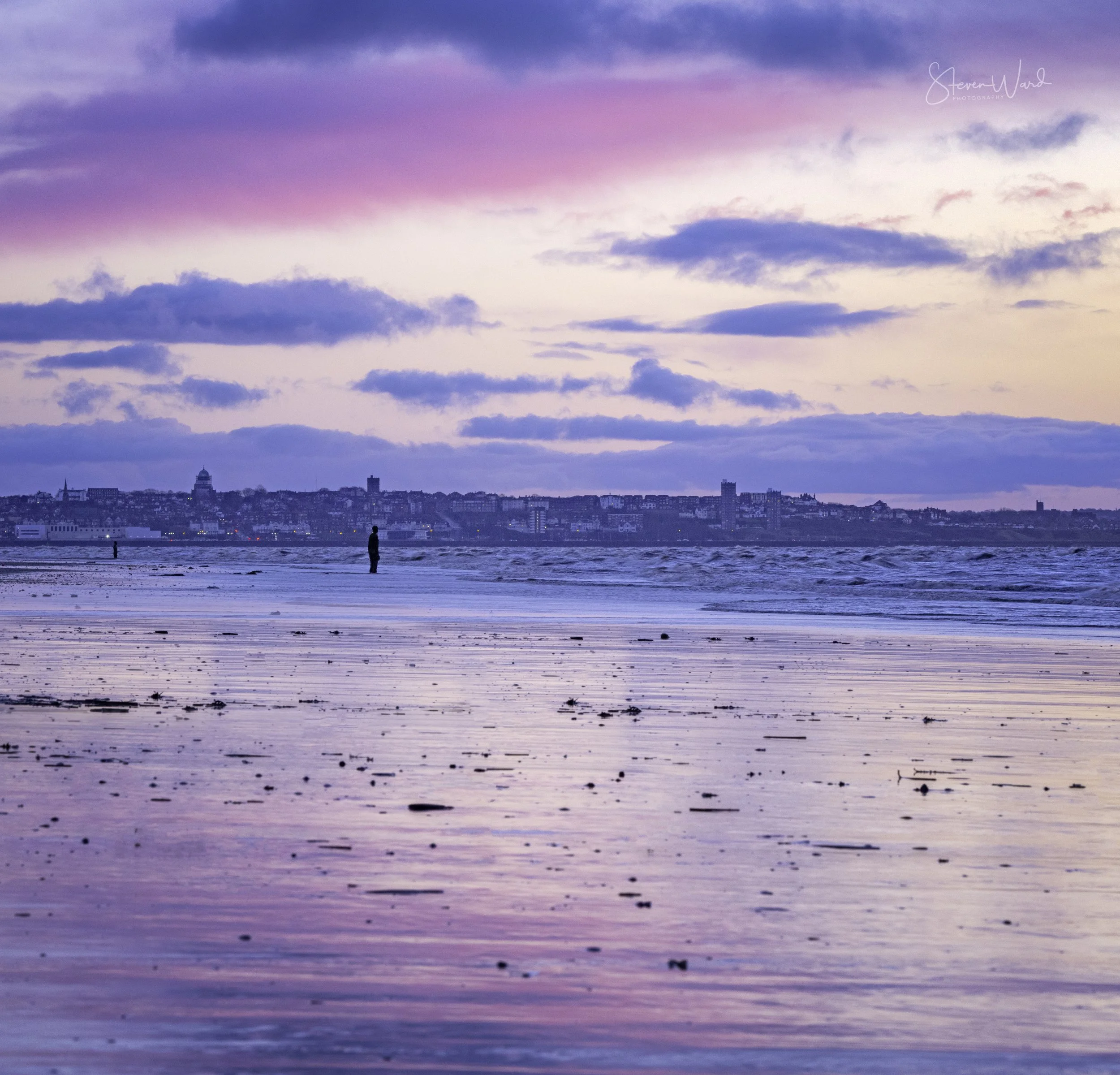 A beach at sunset with pink, purple, and blue sky, reflecting on wet sand, featuring a person standing near the shoreline and a distant city skyline in the background.