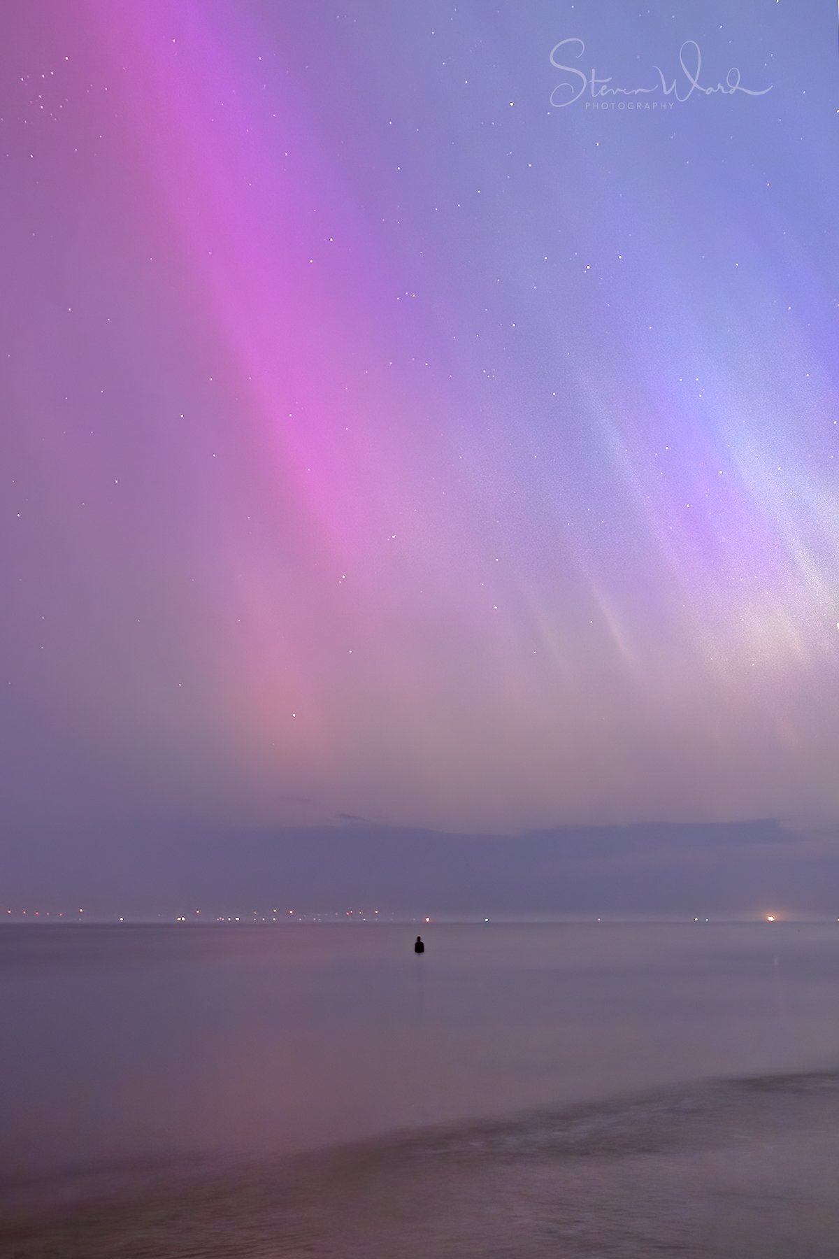 A night sky over the ocean with colorful auroras and stars. A small boat is visible on the water.