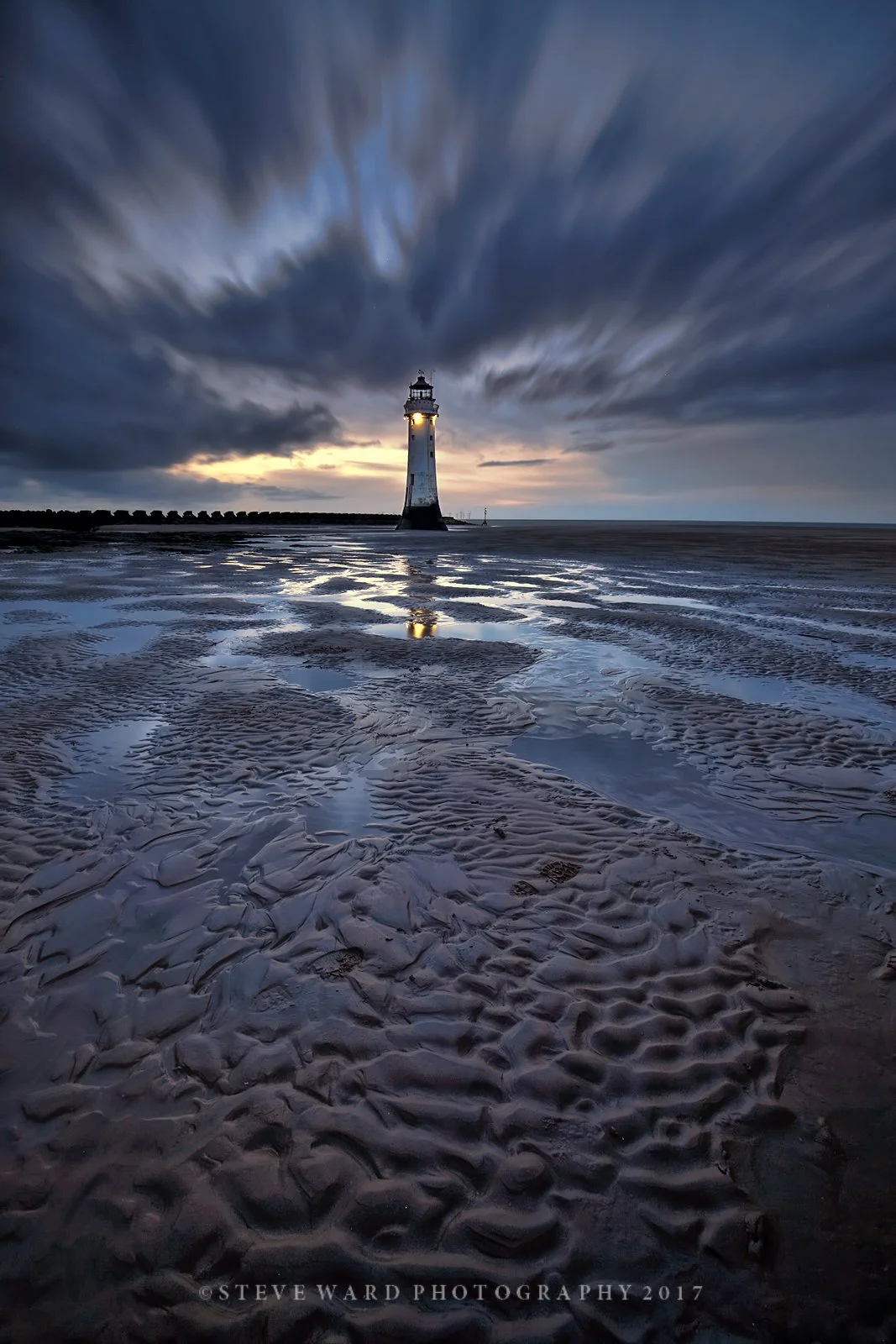 A lighthouse standing on a sandy beach during dusk with dark, cloudy skies and a reflection in the wet sand