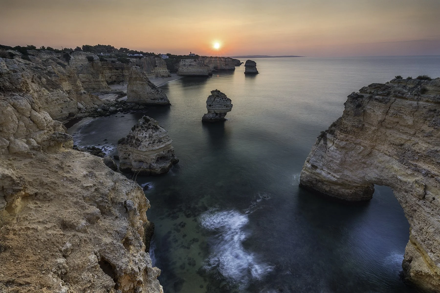 Sunset over rugged cliffs and rock formations along a coastline with calm waters and a small waterfall.