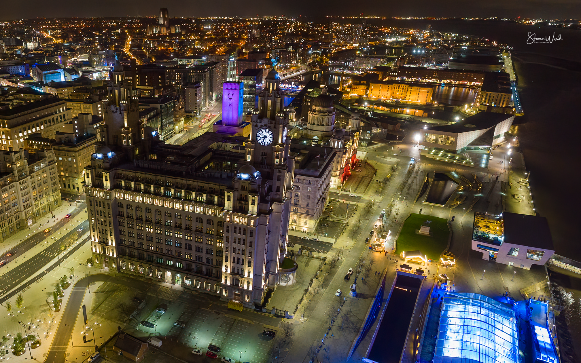 Nighttime aerial view of a cityscape featuring a historic building with a clock tower, modern architecture, and waterfront with illuminated pathways and buildings.