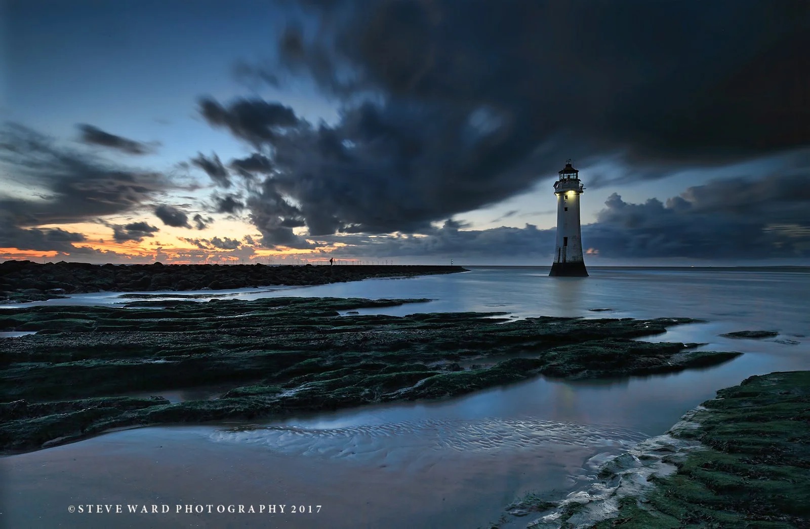 A lighthouse on a rocky shoreline at dusk with dark clouds in the sky and a faint sunset on the horizon.