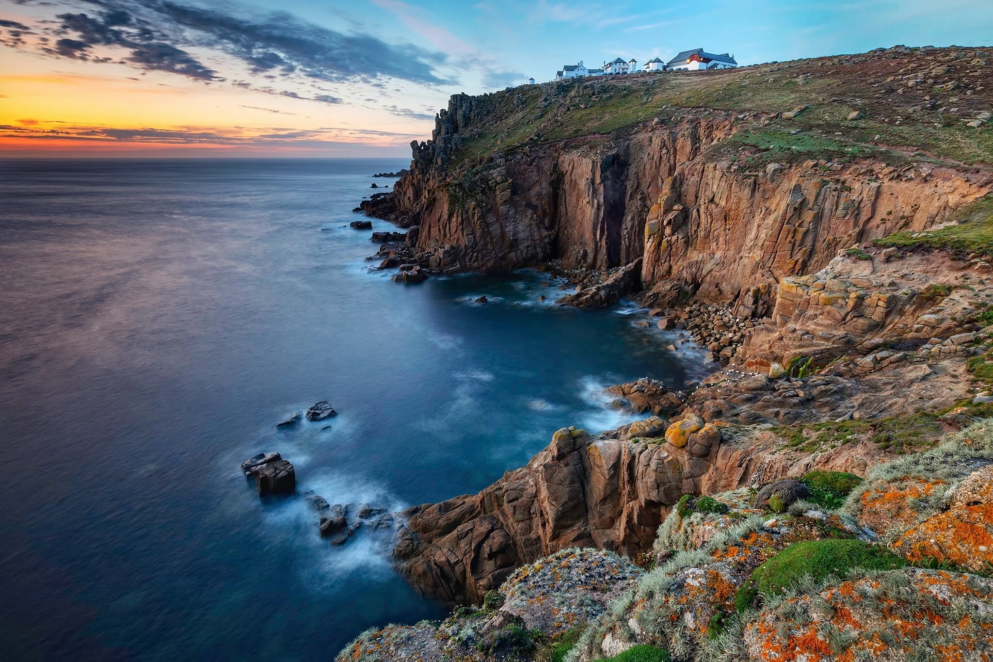 Cliffside coastline with rocky cliffs, ocean waves, and a sunset sky, with buildings on top of the cliffs.