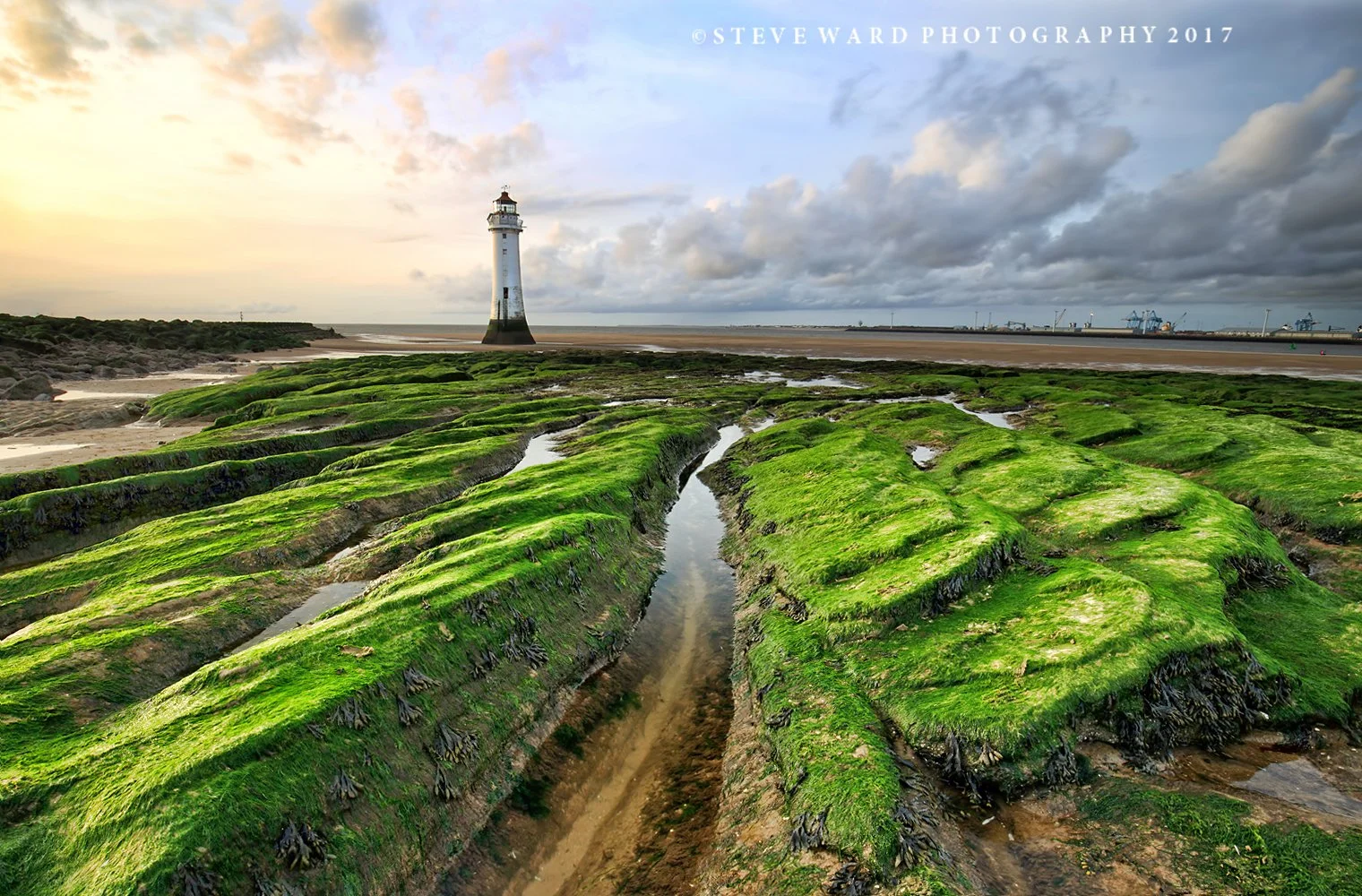 A lighthouse standing on a sandy beach with green moss-covered rocks and tide pools in the foreground, cloudy sky, and a harbor with cranes in the background.