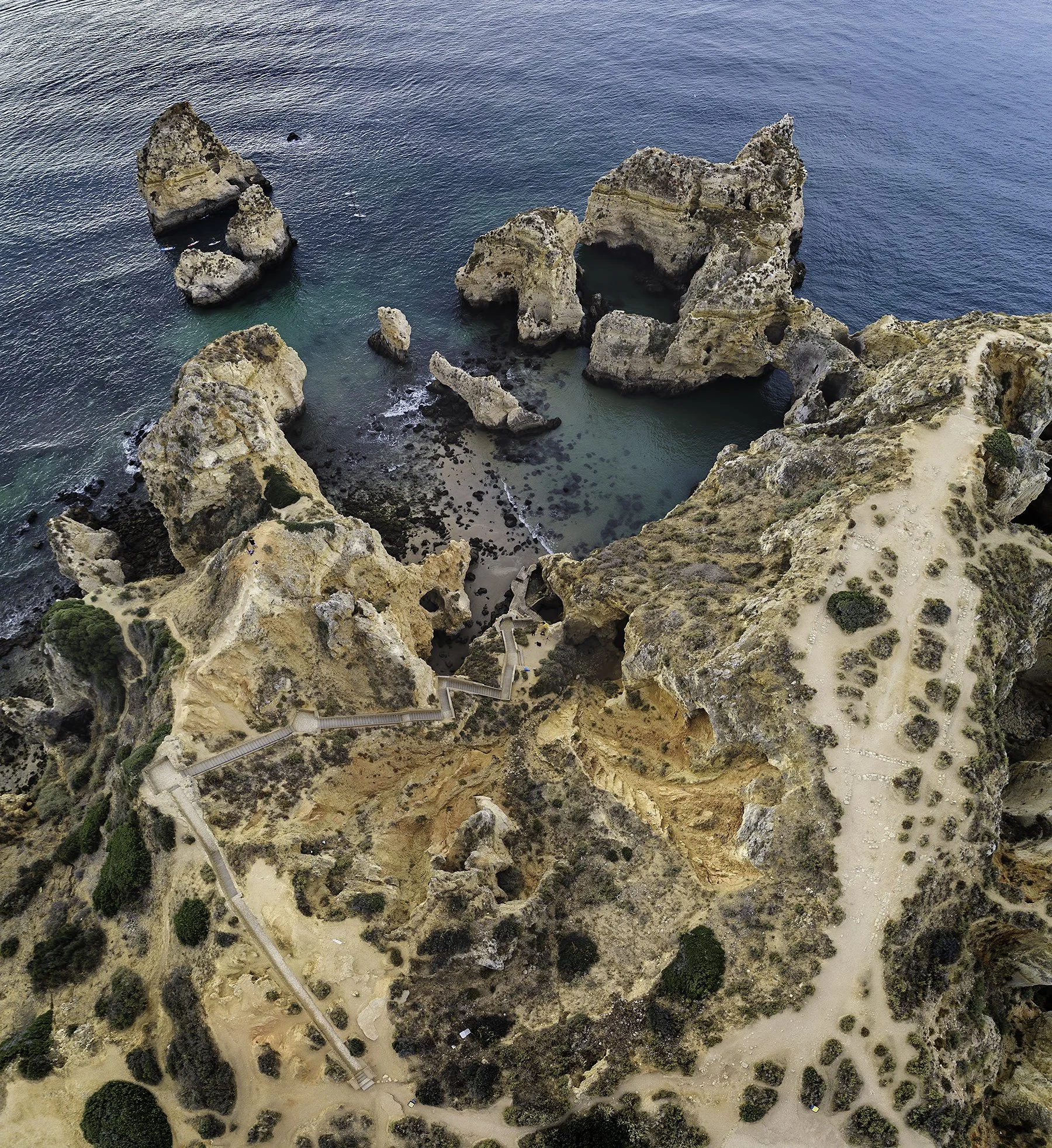 Aerial view of a rugged coastline with large rock formations in turquoise waters, and a sandy walking path along the cliffs with scattered bushes.