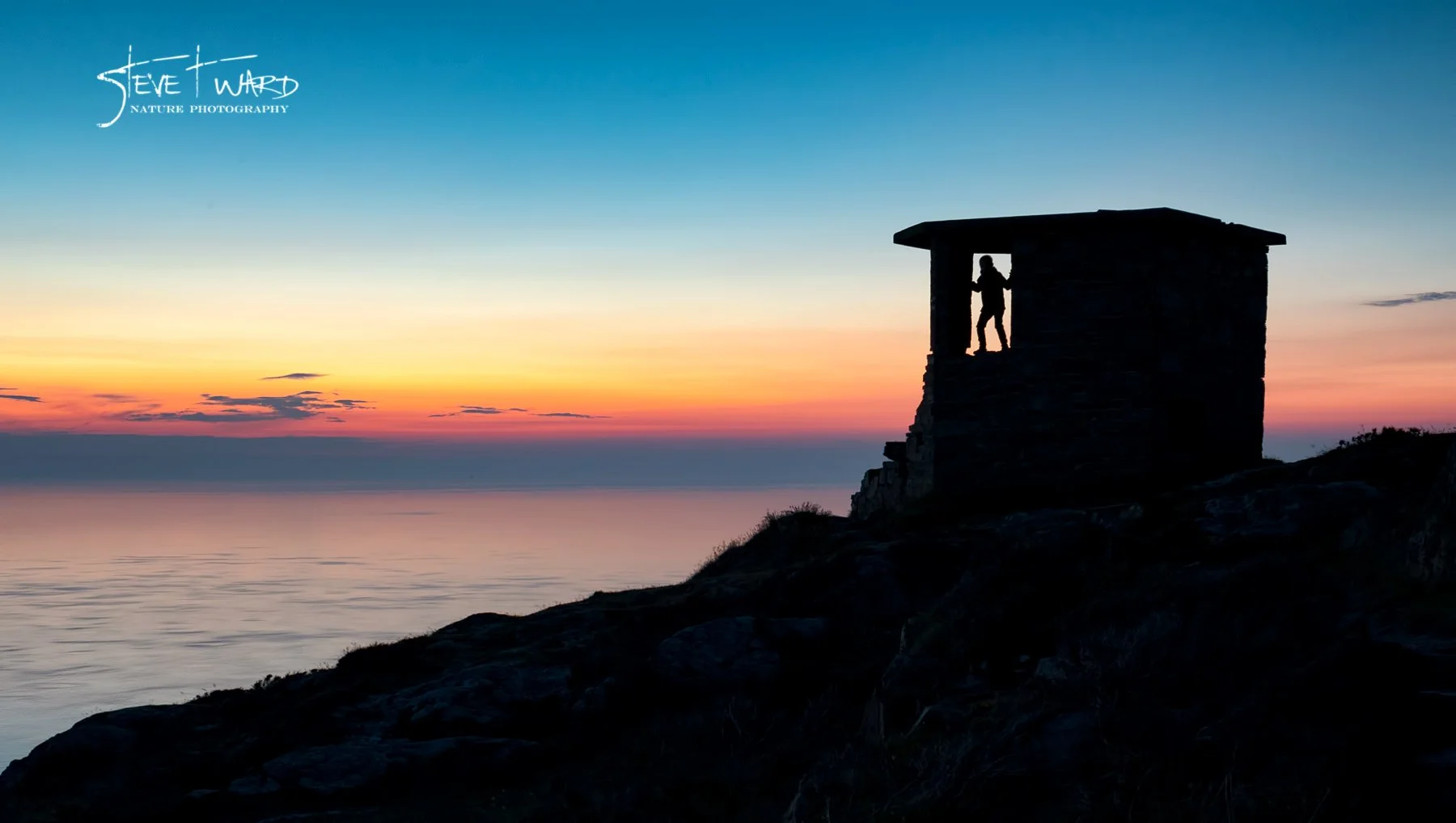 Silhouette of a person standing on a small wooden cabin's balcony at sunset, overlooking the ocean with a colorful sky of pink, orange, and blue hues.