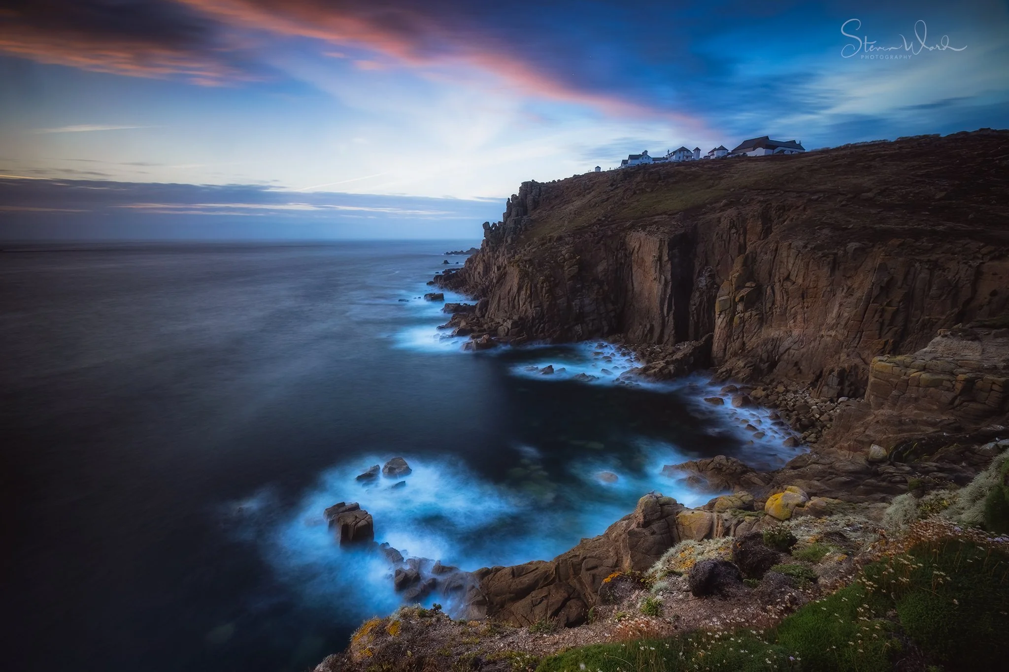 Coastal cliffs with ocean waves crashing against rocks, houses on top of the cliffs, and a colorful sunset sky.