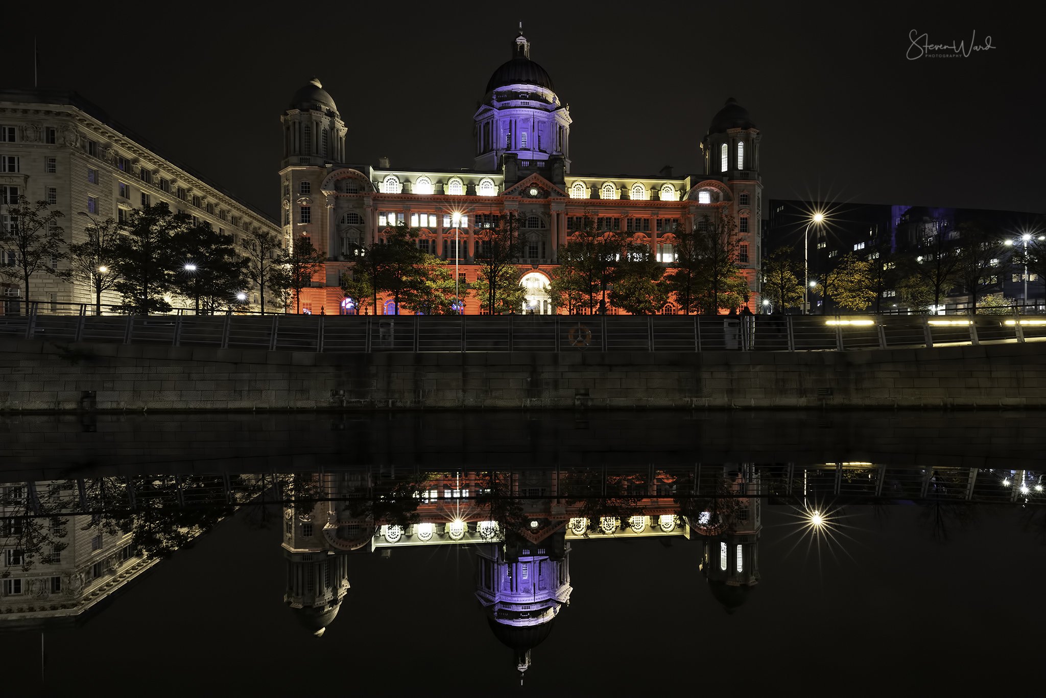 Night view of a historic building with a domed roof, illuminated in red, blue, and white lights, reflected in the water below.