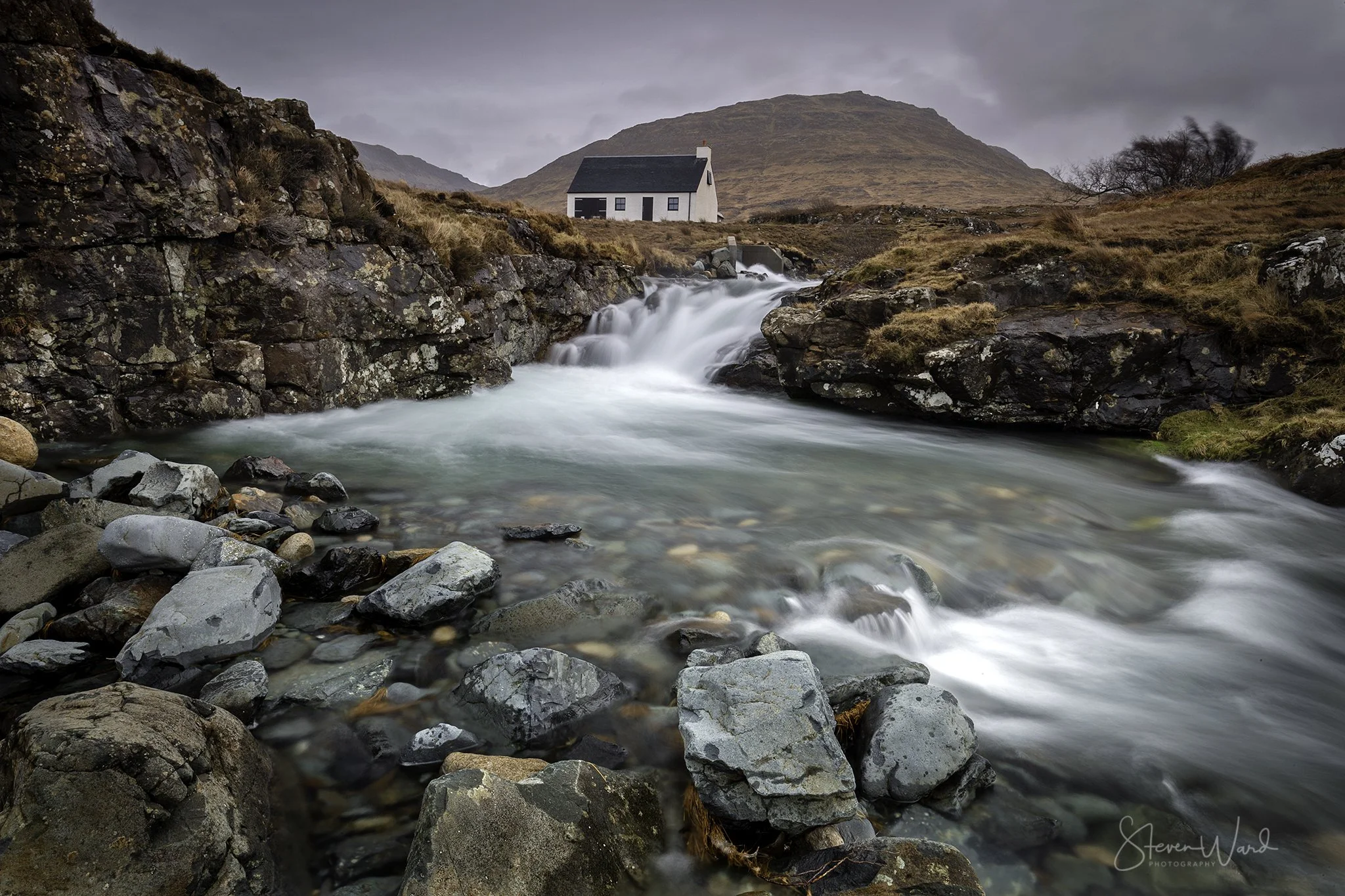 A small river or stream flowing over rocks with a small waterfall, set in a rural landscape with a white house near the river and brown grassy hills in the background under a cloudy sky.