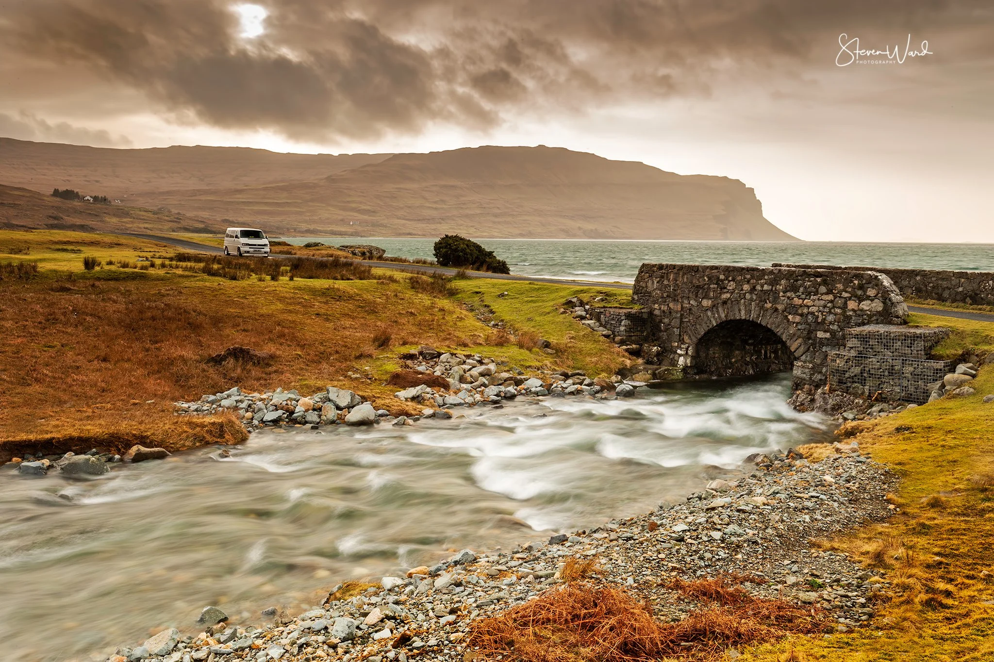 Scenic landscape with a river flowing under a stone bridge, green and brown grassy banks, a vehicle driving on a nearby road, distant hills, cloudy sky, and ocean in the background.