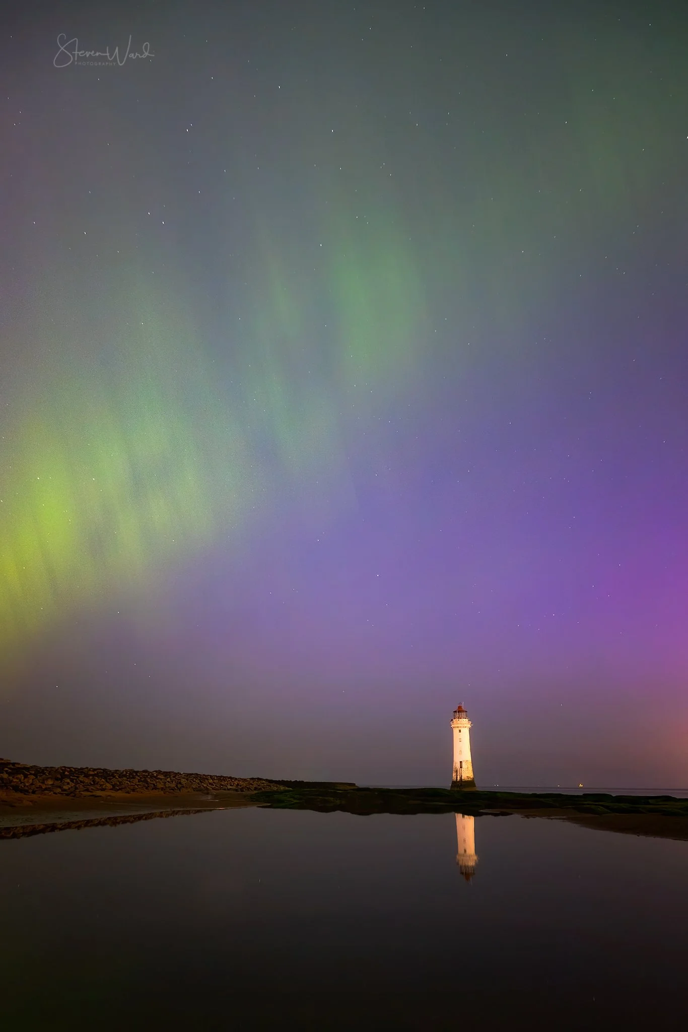 Night sky with colorful northern lights and stars, lighthouse on the coastline reflected in a calm body of water.