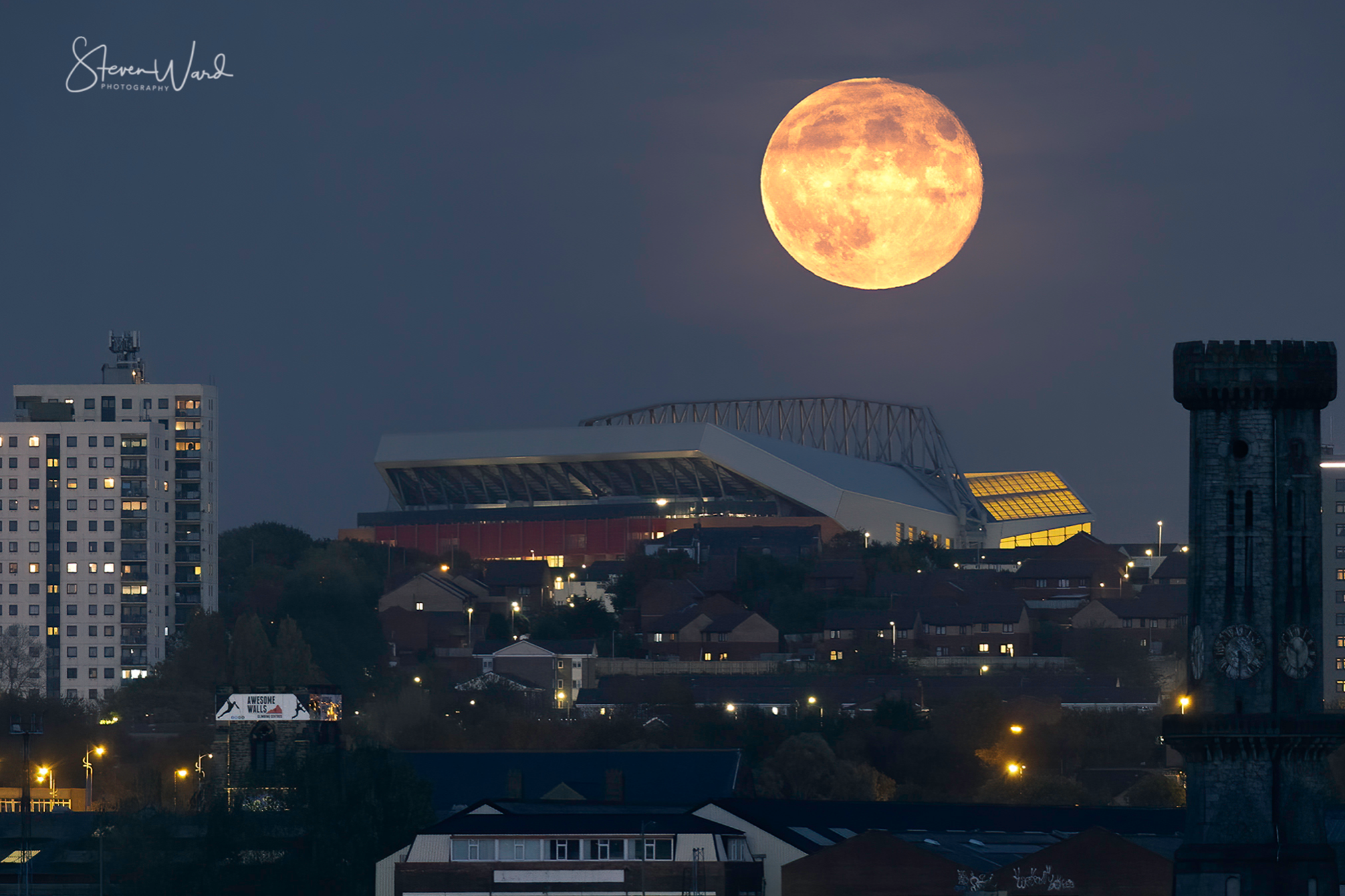 Large, full moon rising over a city skyline at night with lit windows and streetlights, including a sports stadium and a clock tower.
