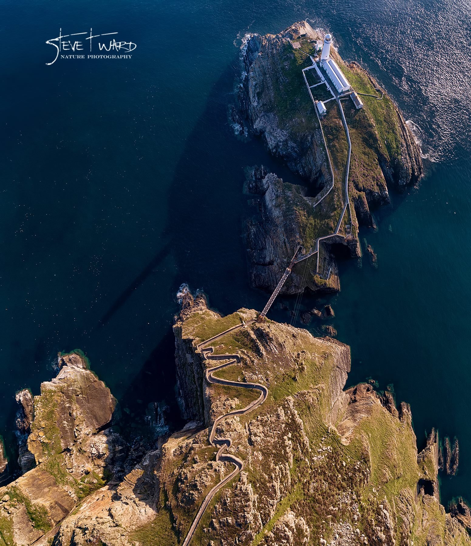 Aerial view of an island with a lighthouse on top, connected by a winding path and a bridge, surrounded by dark blue ocean water.