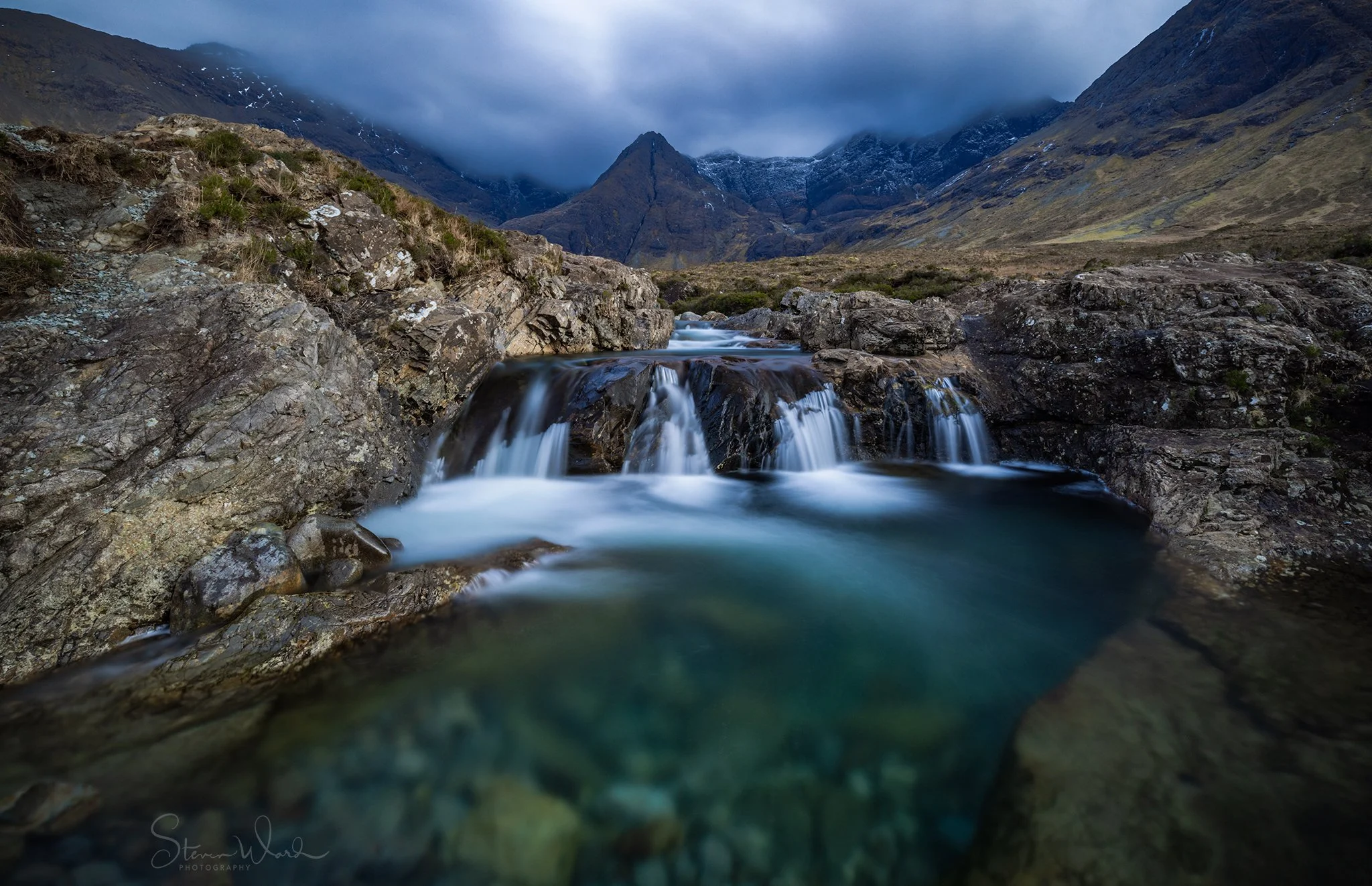 Scenic mountain landscape with a small waterfall flowing over rocks into a clear river, mountains shrouded in clouds in the background.